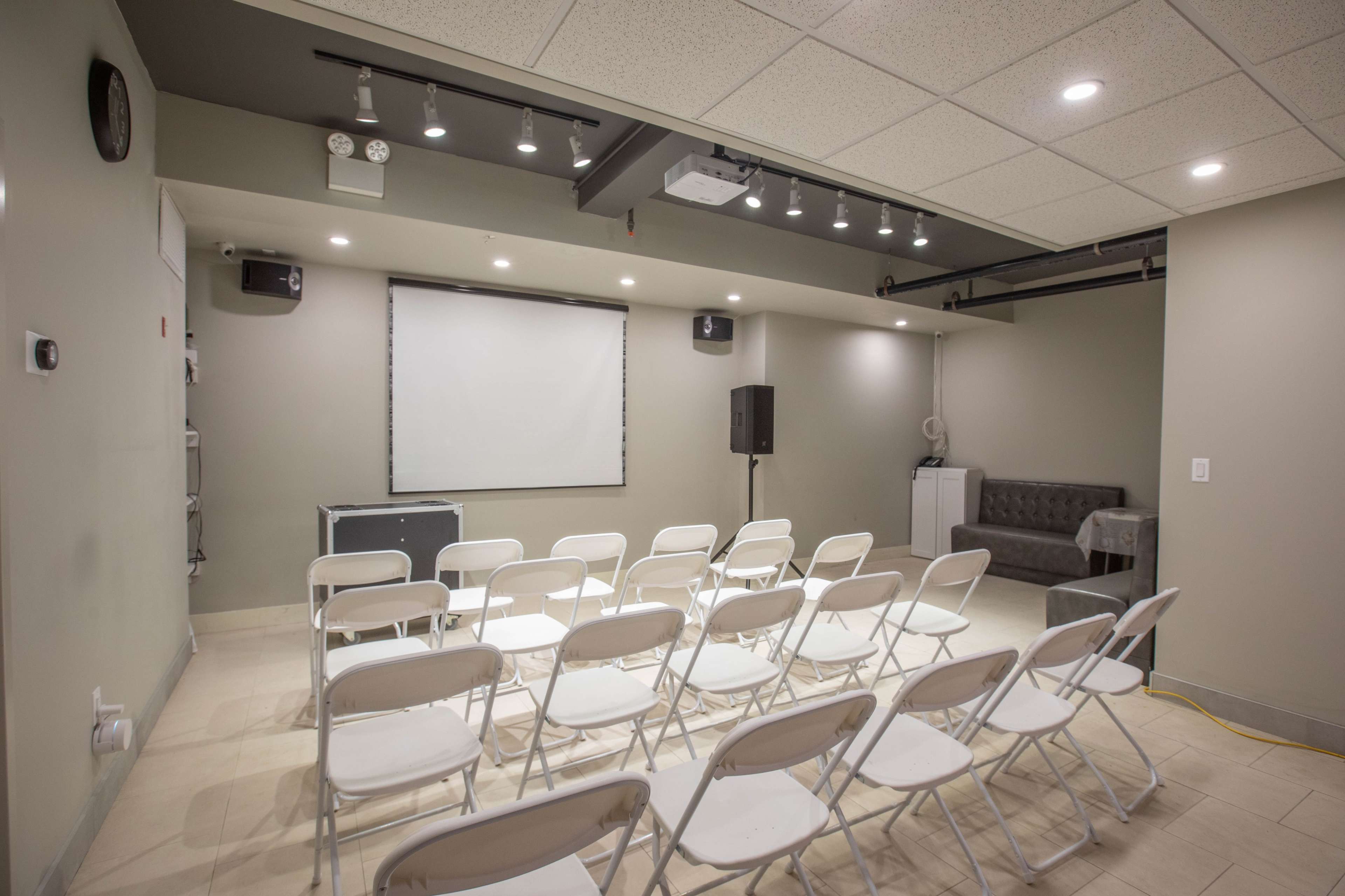 A seminar room with rows of white chairs facing a blank projection screen and a sound system in the corner.