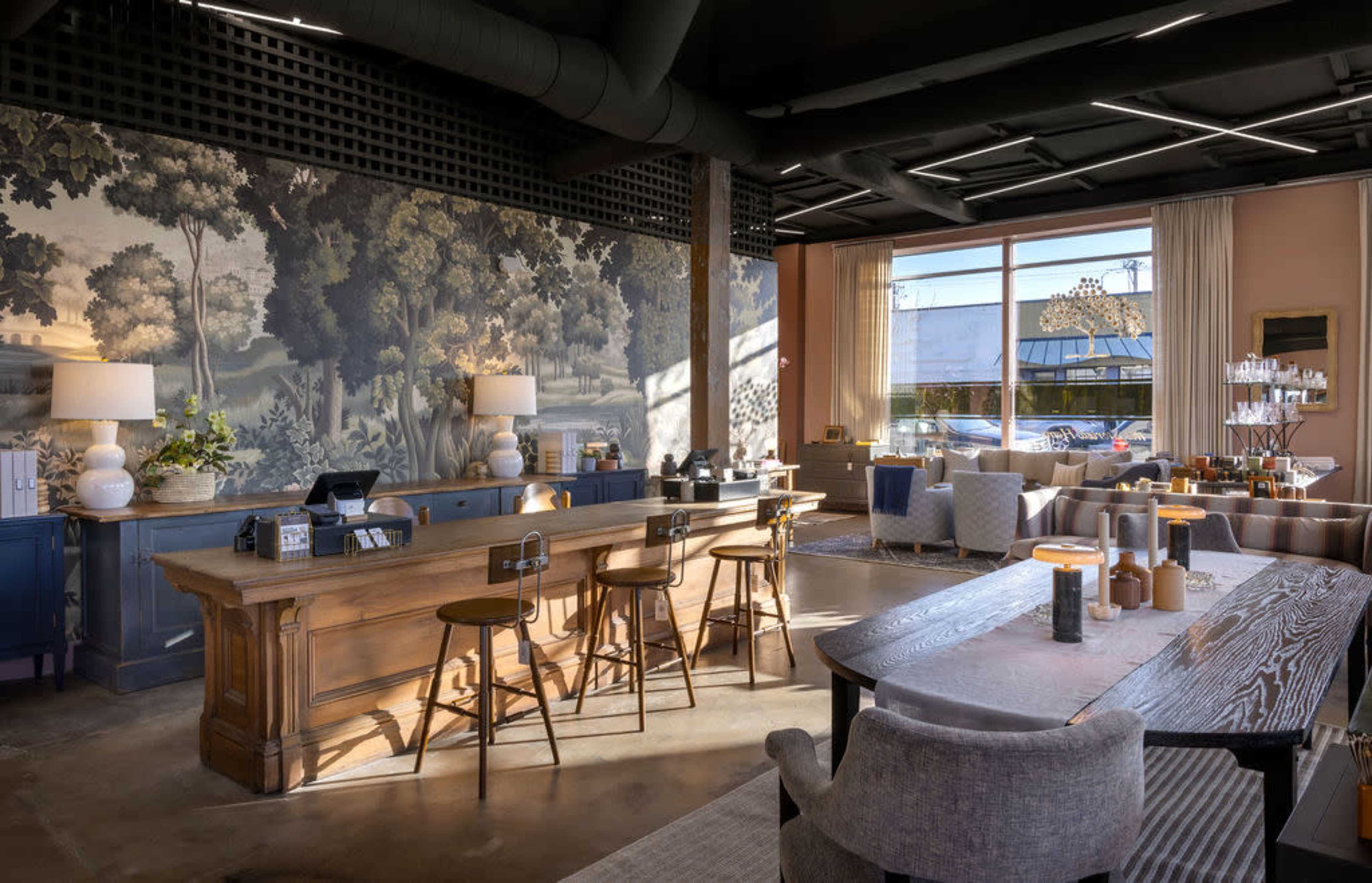 A modern interior of a café or lounge with a wooden counter, seating, and large windows illuminating a mural of trees on the wall.