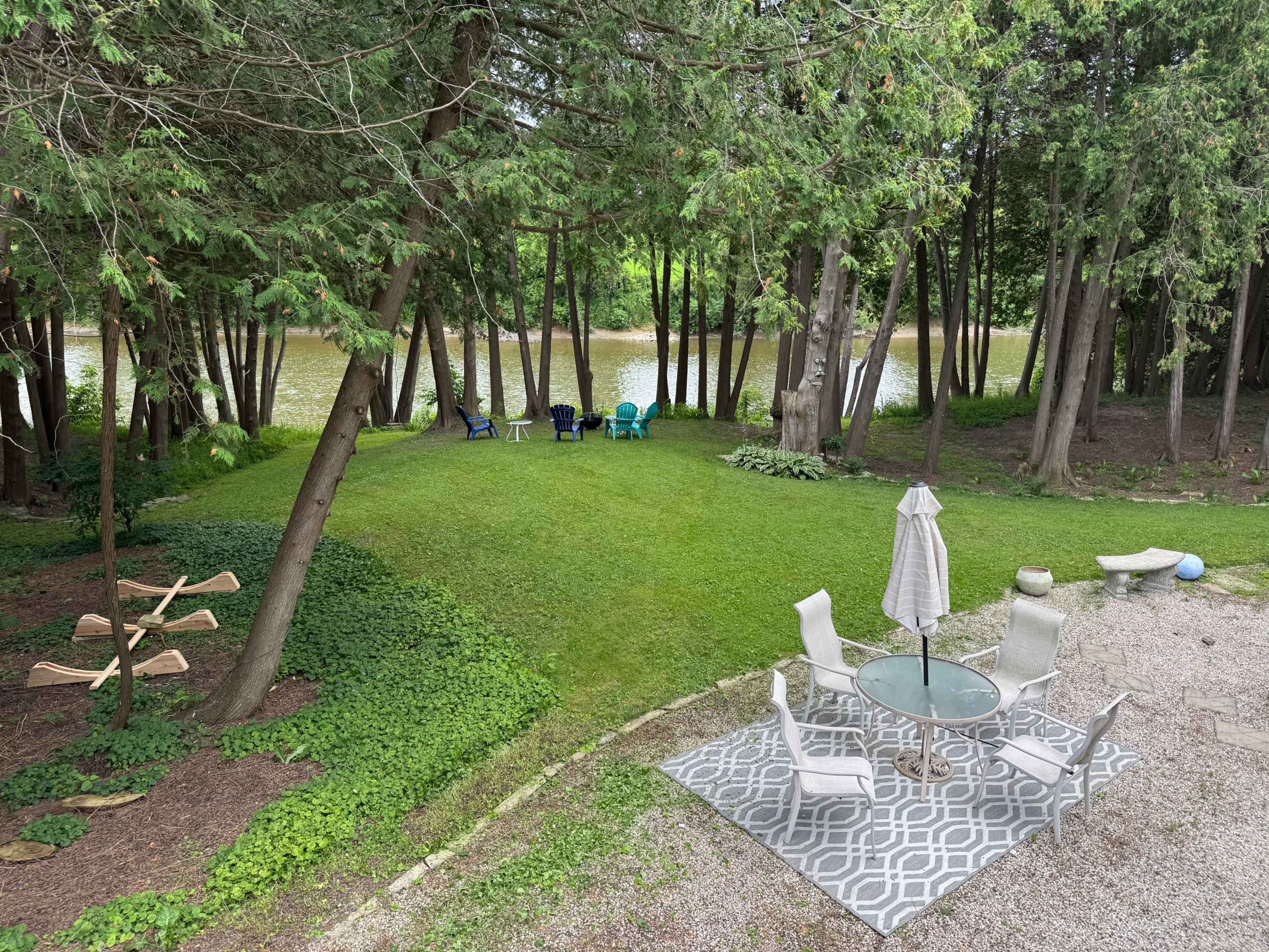 The image shows a grassy backyard with a patio table and chairs, a row of trees, and a river flowing in the background.