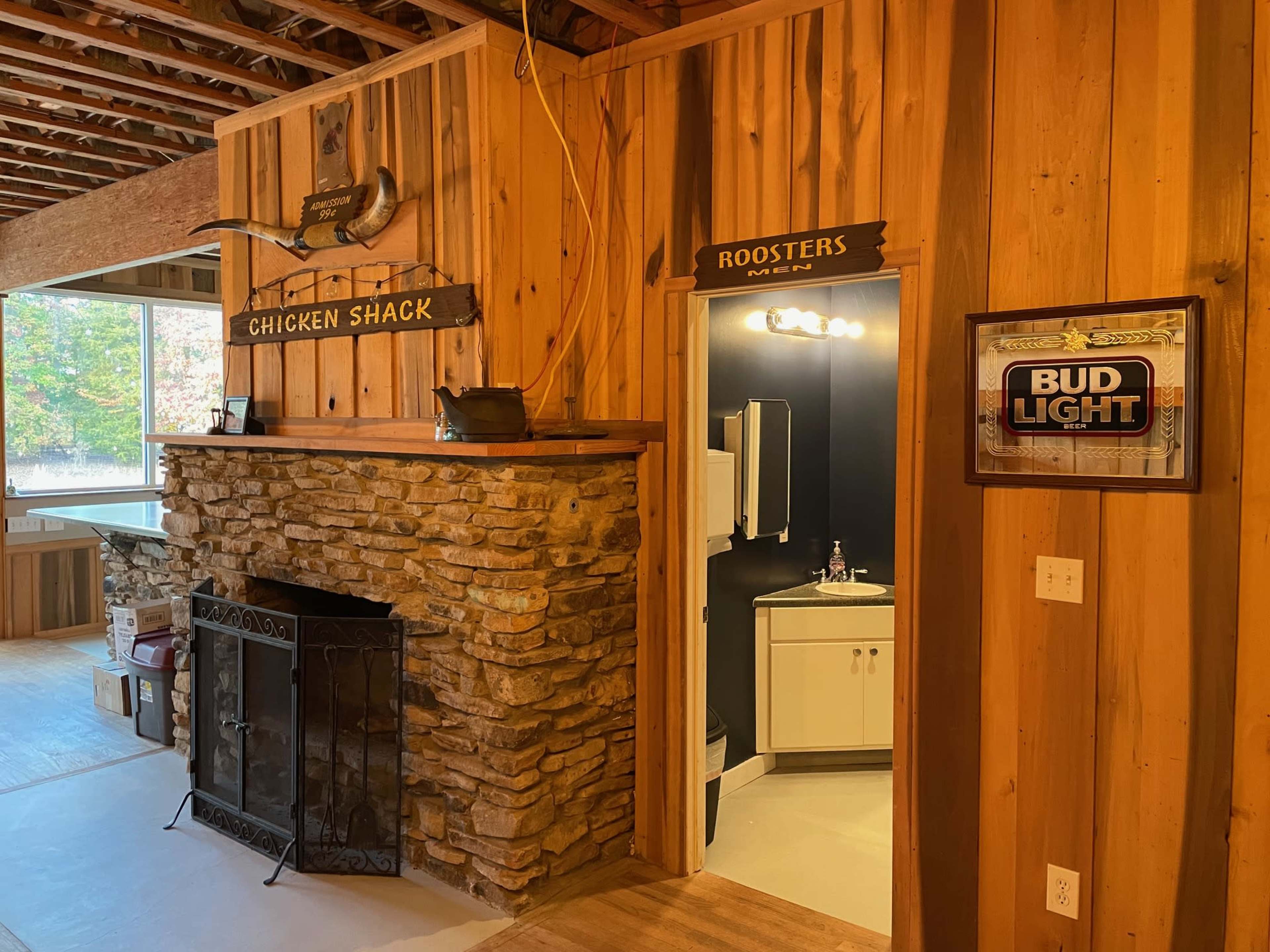 A rustic interior with wooden walls, featuring a stone fireplace, signs labeled "CHICKEN SHACK" and "ROOSTERS," and a doorway leading to a bathroom.