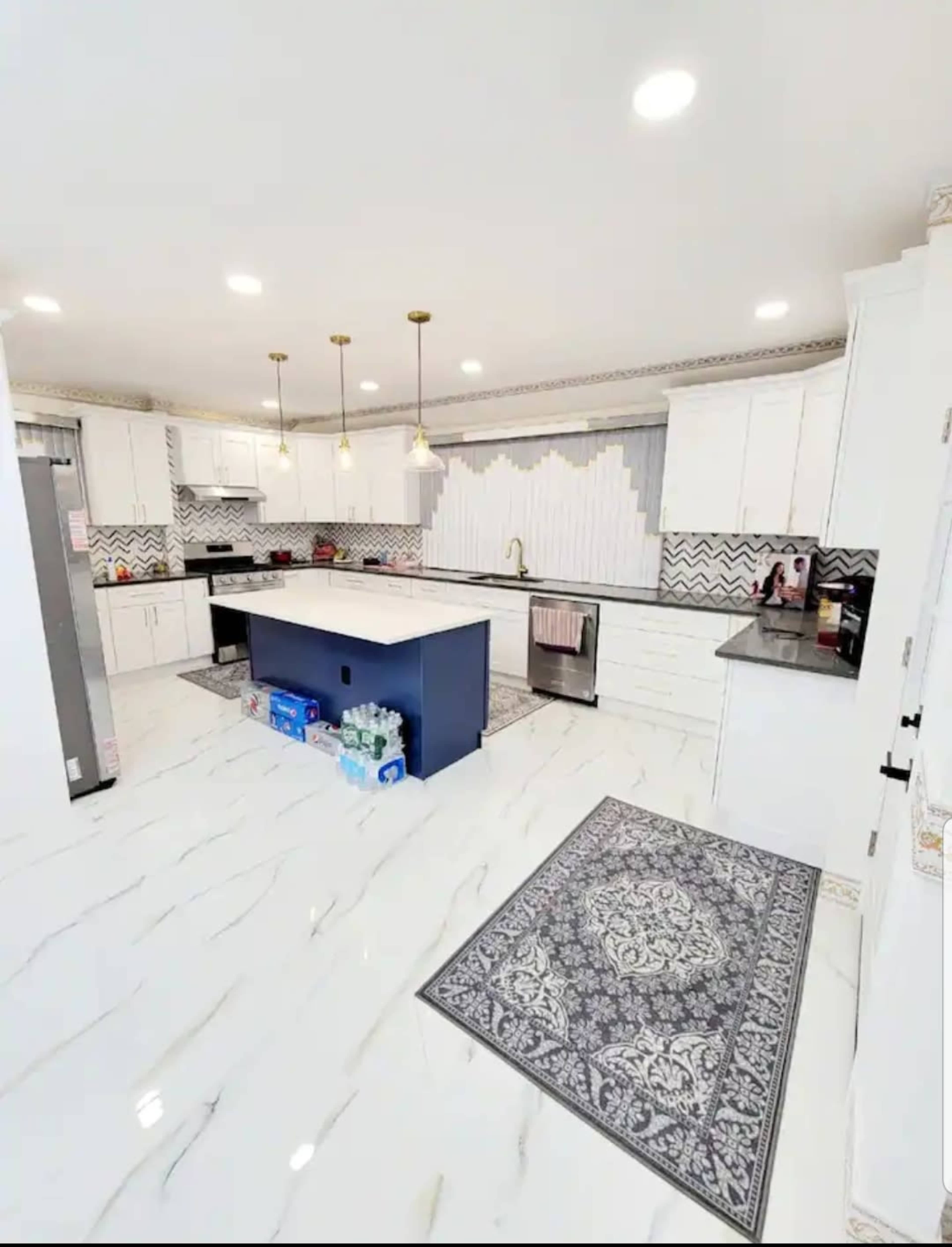 A modern kitchen features white cabinets, a blue island, and a patterned area rug on the marble tile floor.