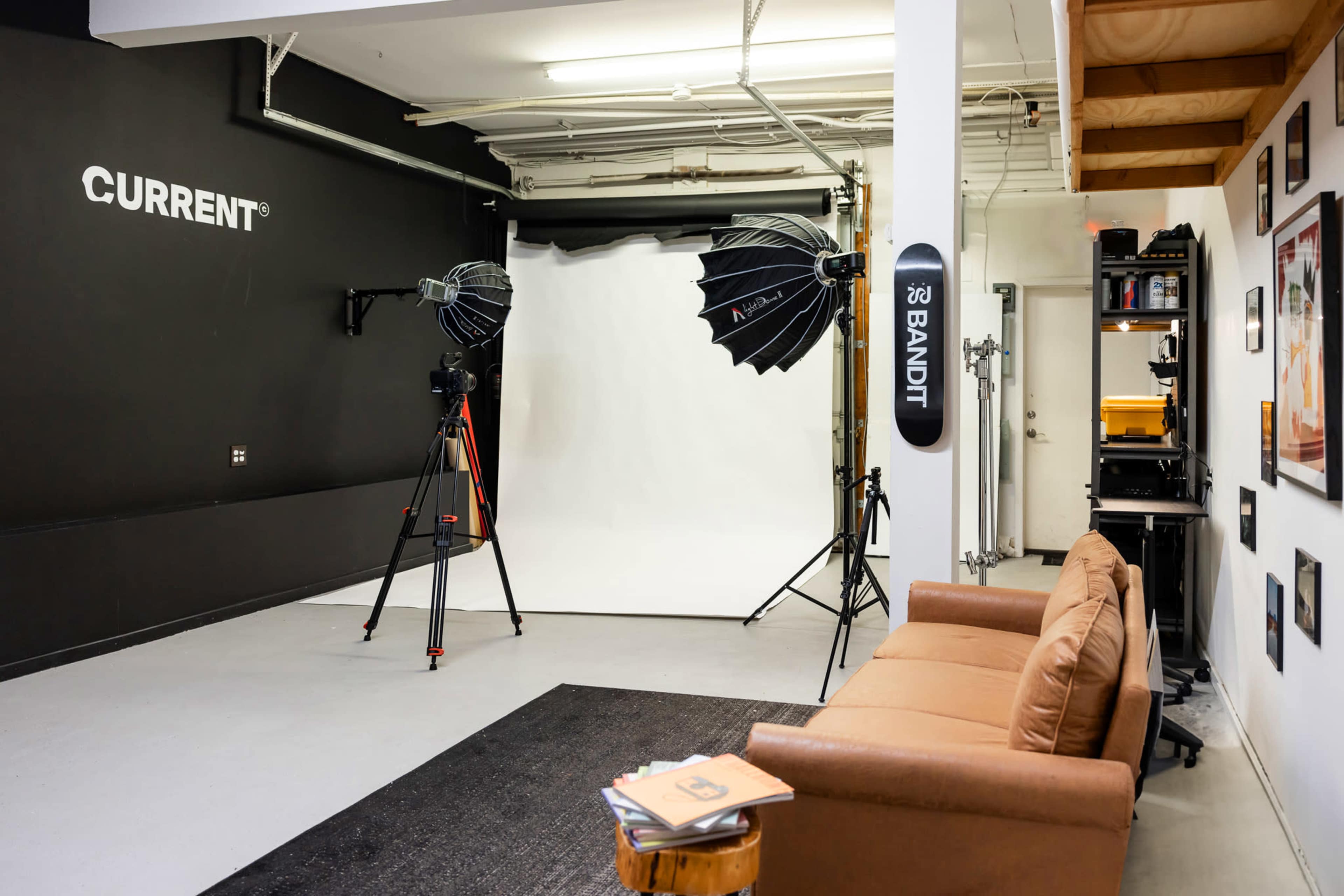 A photography studio with a black wall, a white backdrop, two softbox lights on tripods, a brown couch, and a shelf against the wall filled with equipment.