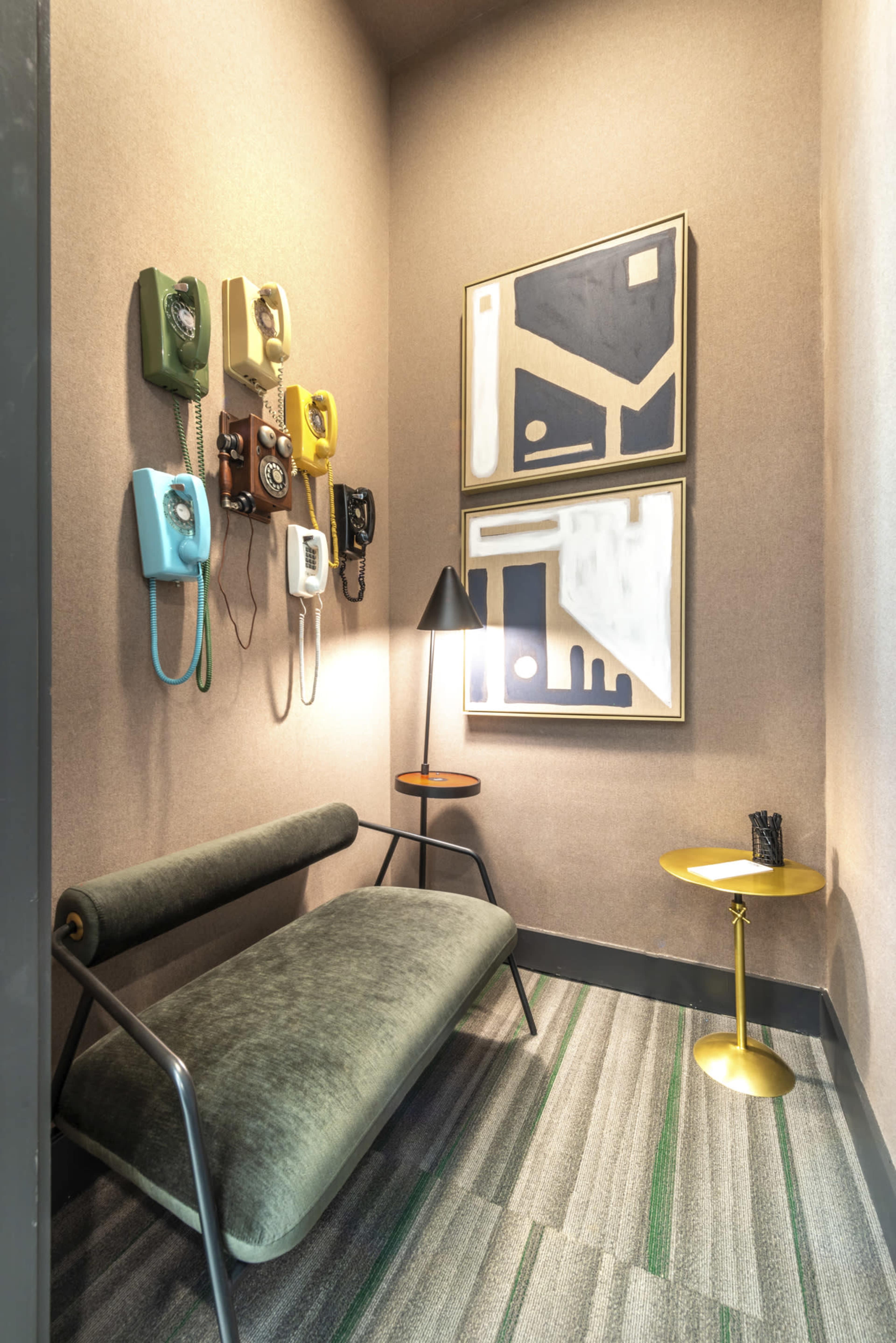 A narrow corner of a room features a green couch, a small round table, a floor lamp, and several vintage telephones mounted on the wall alongside abstract artwork.