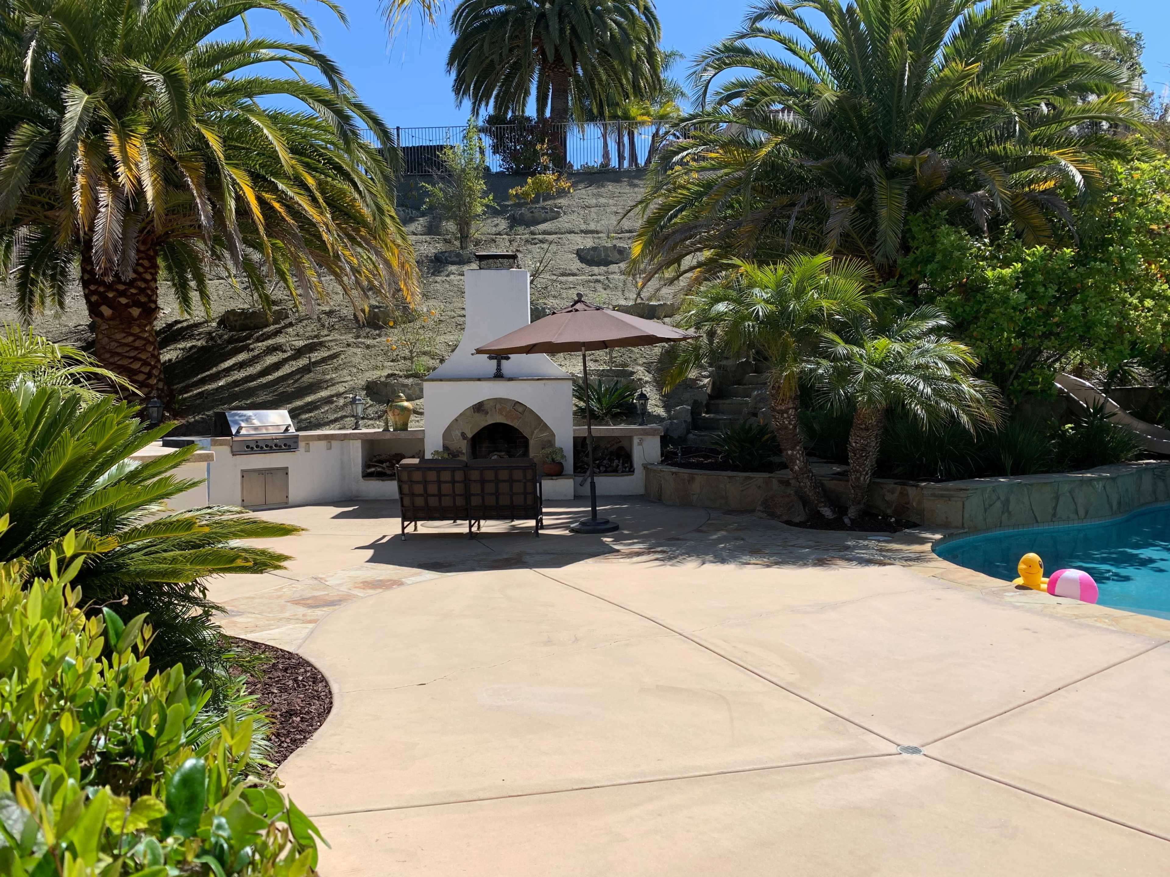 A backyard with a swimming pool, a shaded seating area under an umbrella, a stone fireplace, and palm trees in the background.