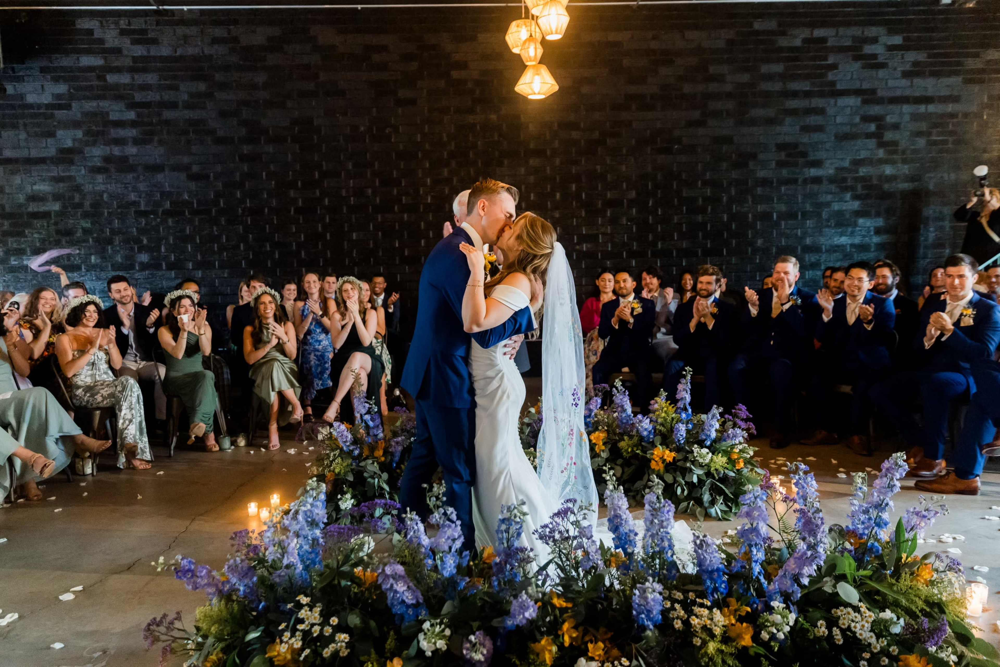 A couple is sharing a kiss at their wedding ceremony, surrounded by floral arrangements and an audience of seated guests applauding.
