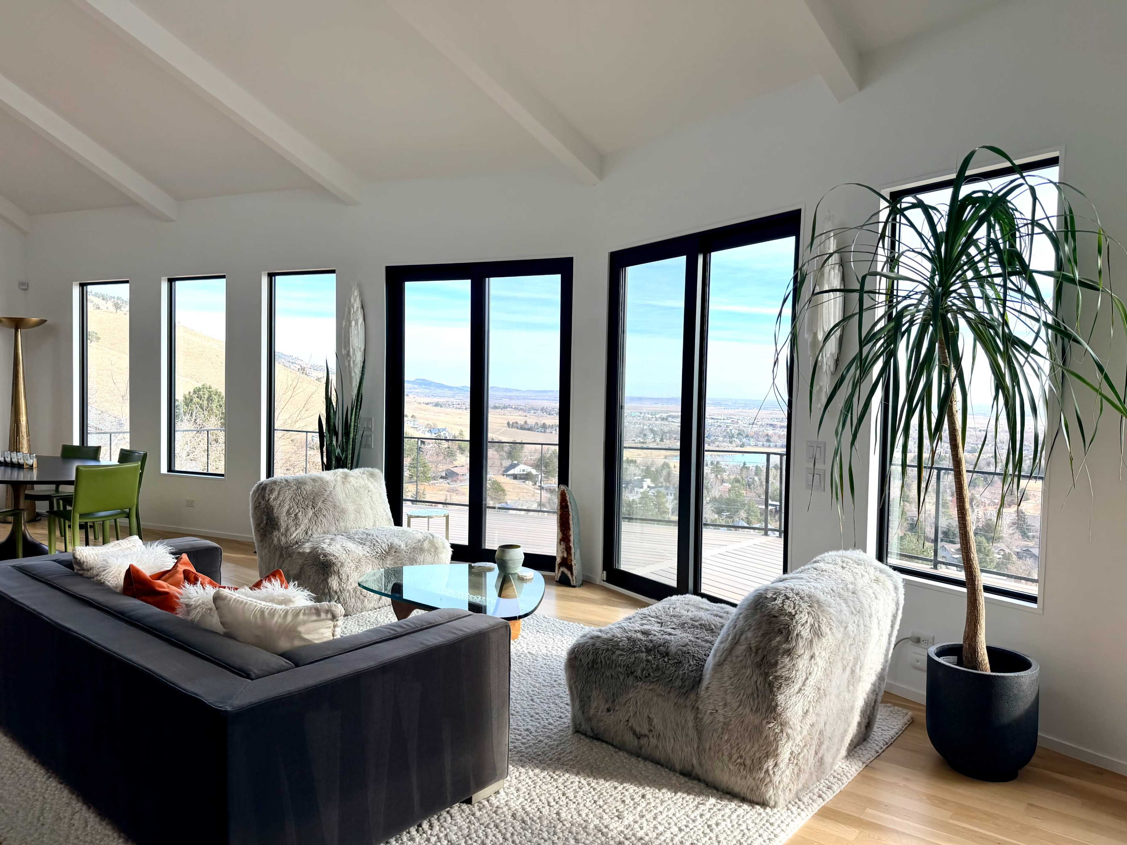 The image shows a modern living room with large windows offering a view of a landscape, featuring a dark sofa, two plush chairs, and a potted plant.