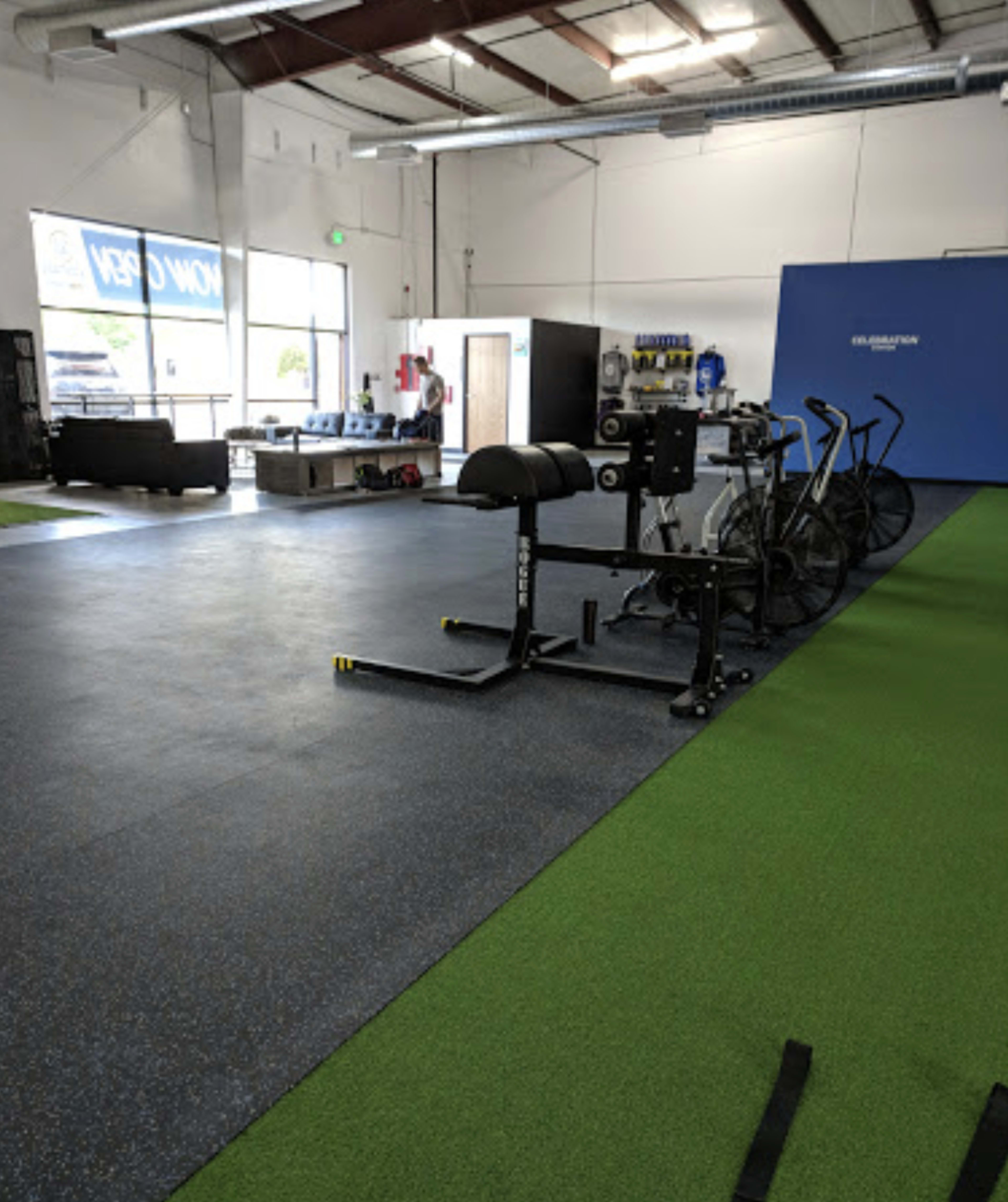 The image shows the interior of a fitness facility with exercise equipment, including a bike and a bench, positioned on a turf-covered floor.