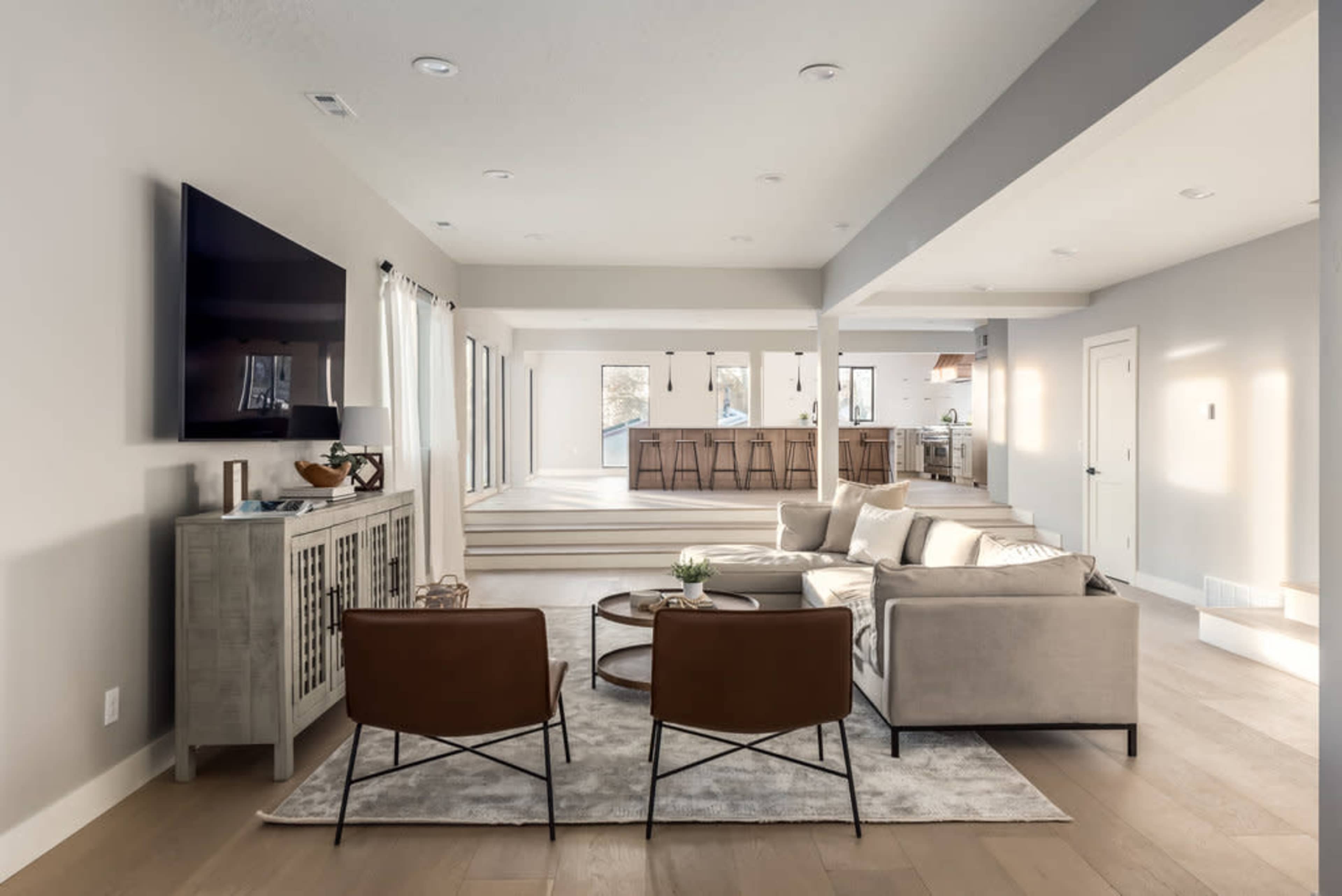 A modern living room with two brown chairs facing a beige sofa and a wall-mounted television, leading into an open-concept kitchen area.