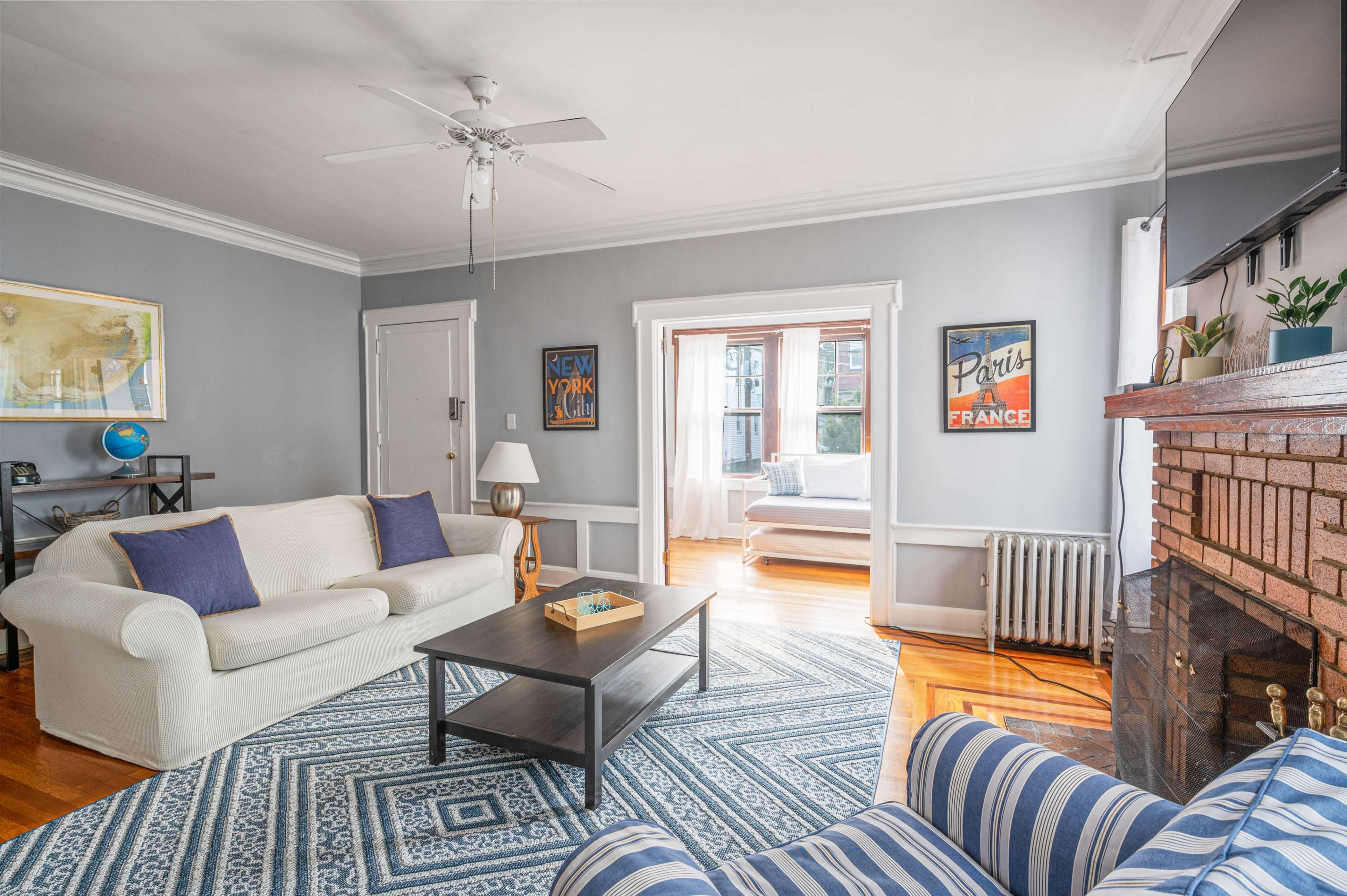 The living room features a light-colored sofa, a blue-striped armchair, a coffee table, and a brick fireplace, with large windows allowing natural light to flood the space.