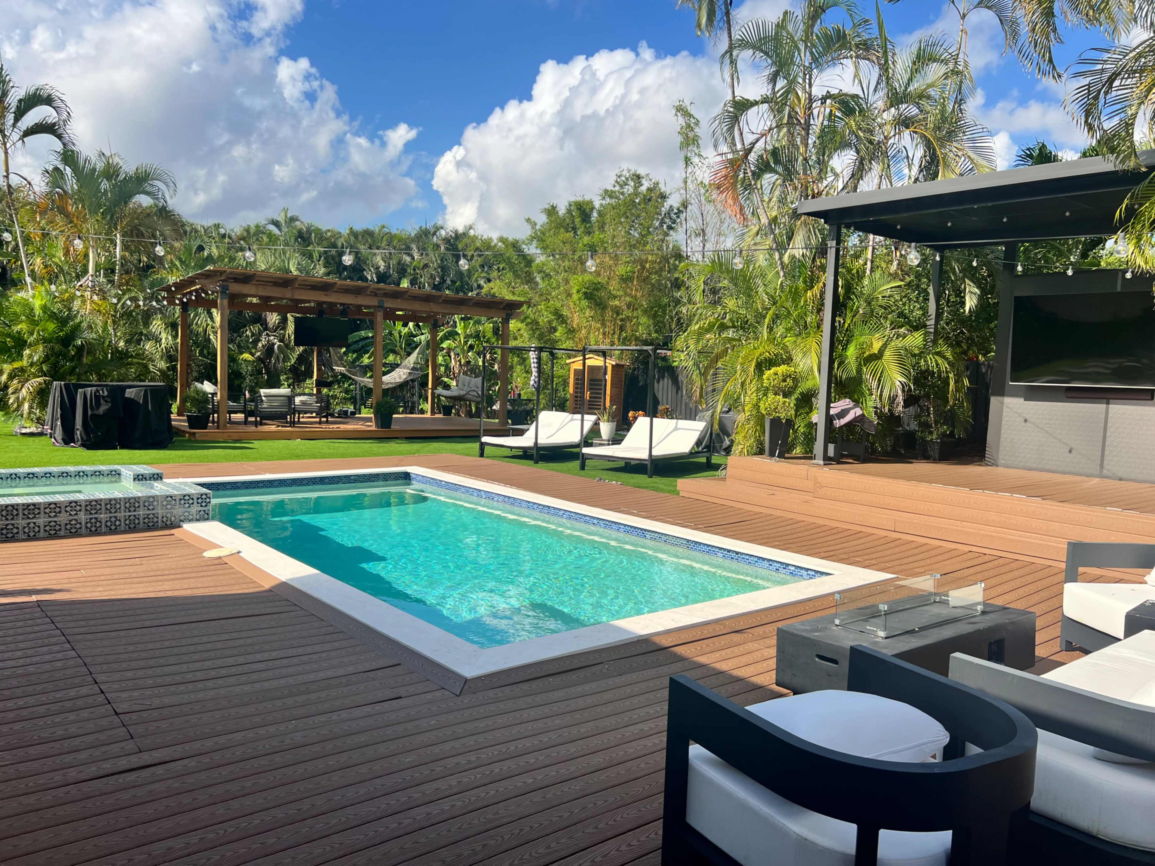 The outdoor space features a swimming pool surrounded by wooden decking, lounge chairs, and a pergola, set amidst tropical greenery.