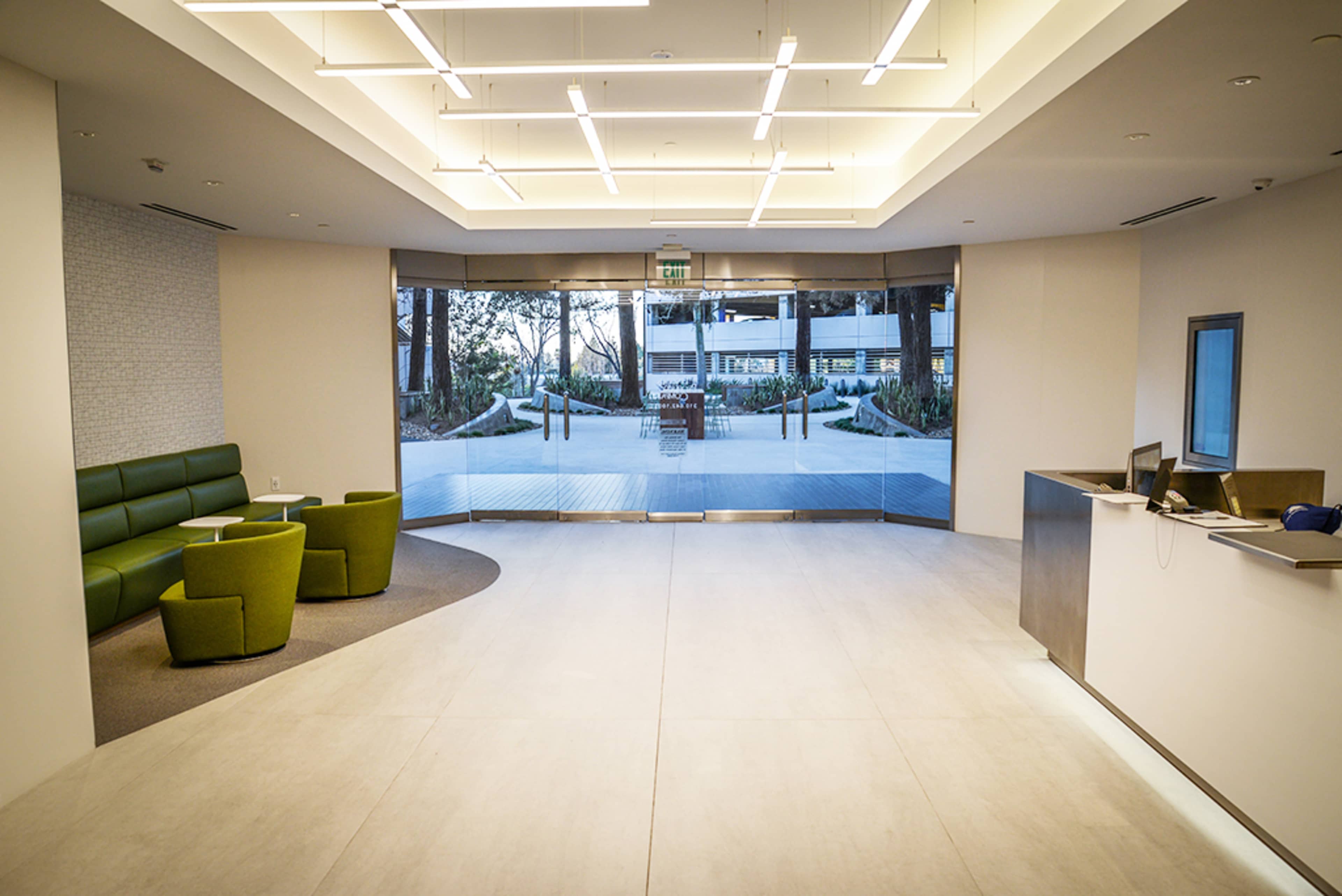 The image shows a modern reception area with a green seating arrangement, a light-colored floor, and a large window displaying an outdoor landscape.