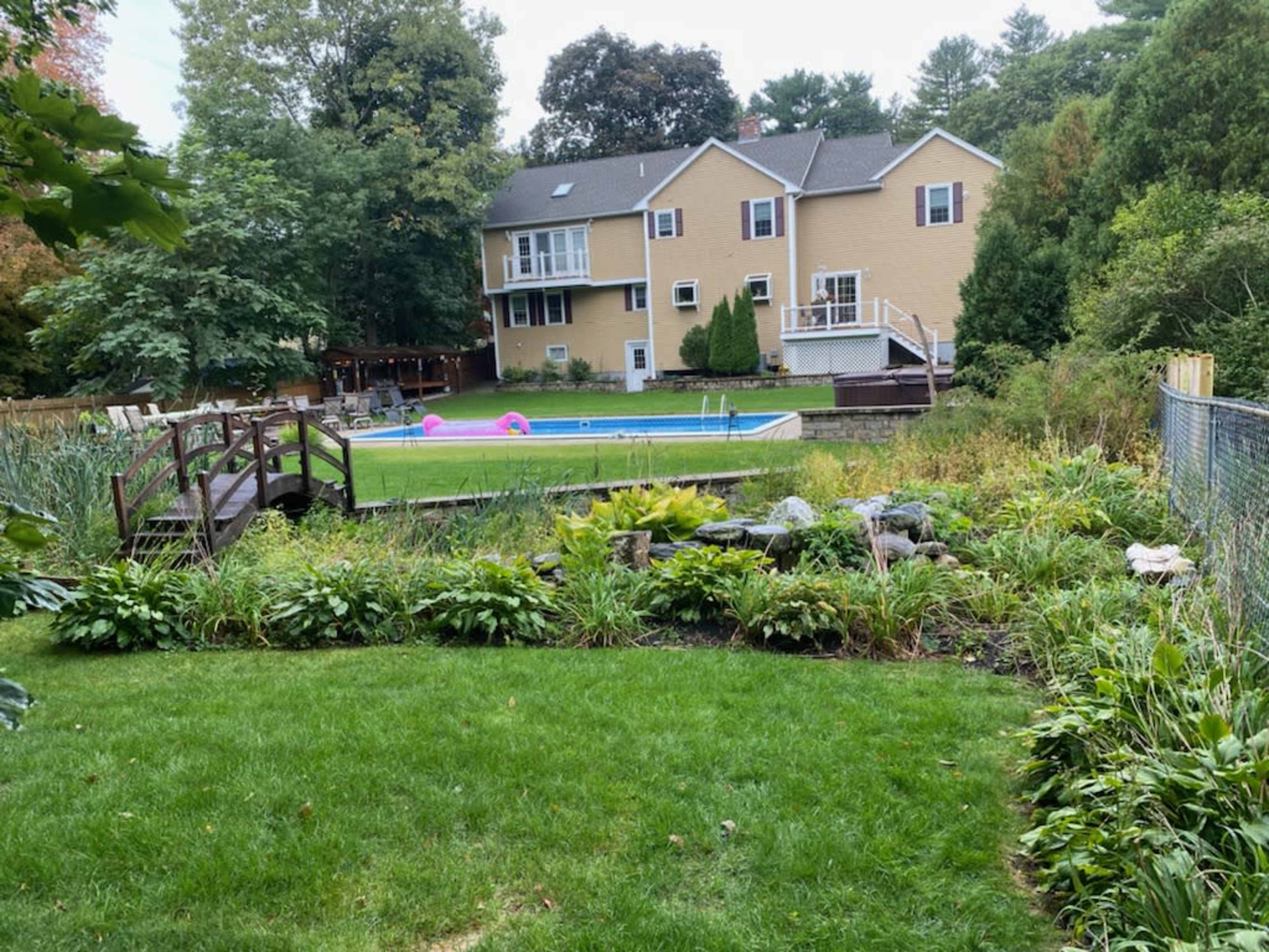 A backyard with a swimming pool, surrounded by landscaping and a wooden bridge leading to an area with more vegetation and trees.