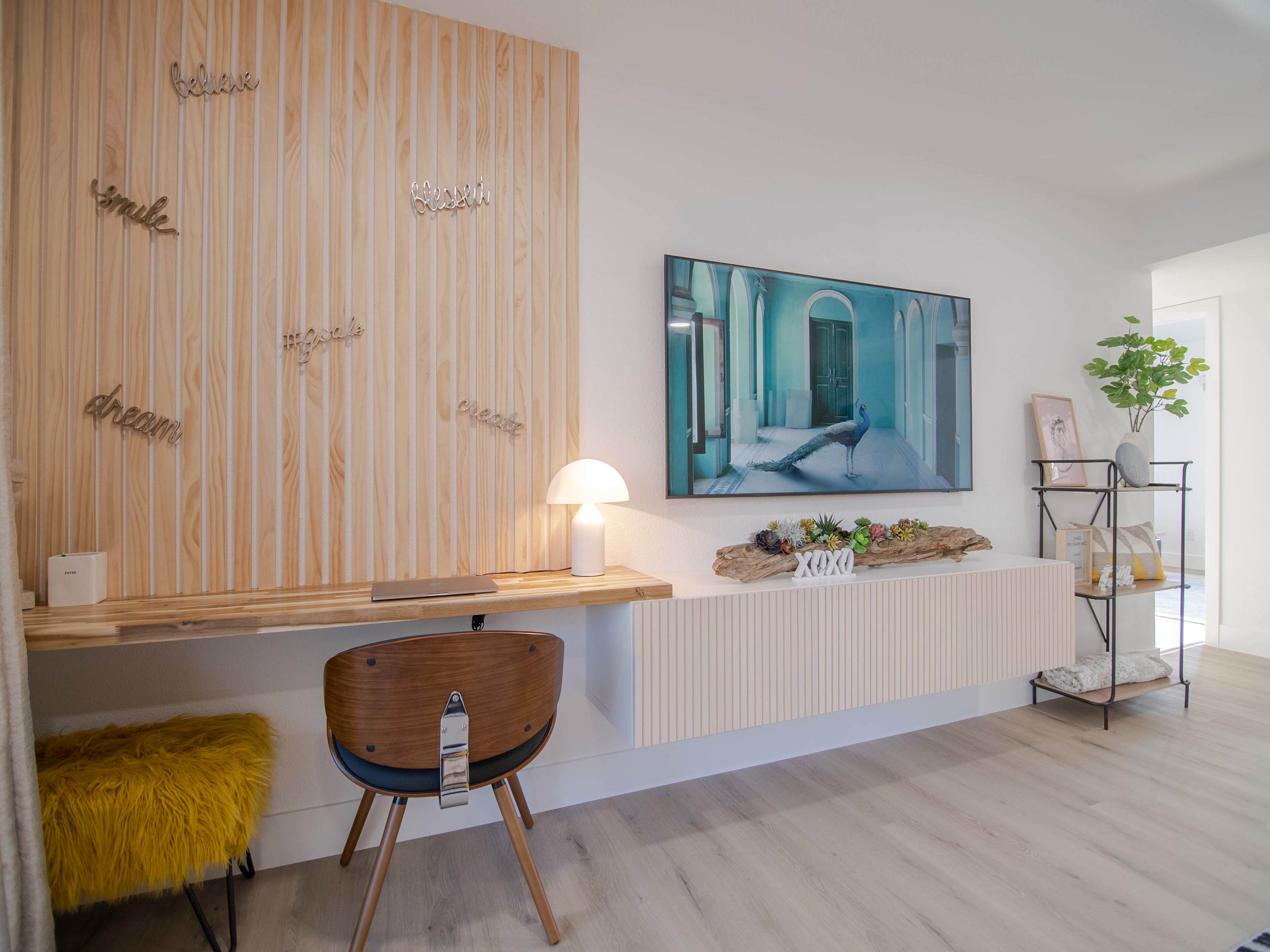 A modern interior space featuring a wooden wall with inspirational words, a desk with a lamp, a TV mounted on the wall, and a decorative shelf with plants.