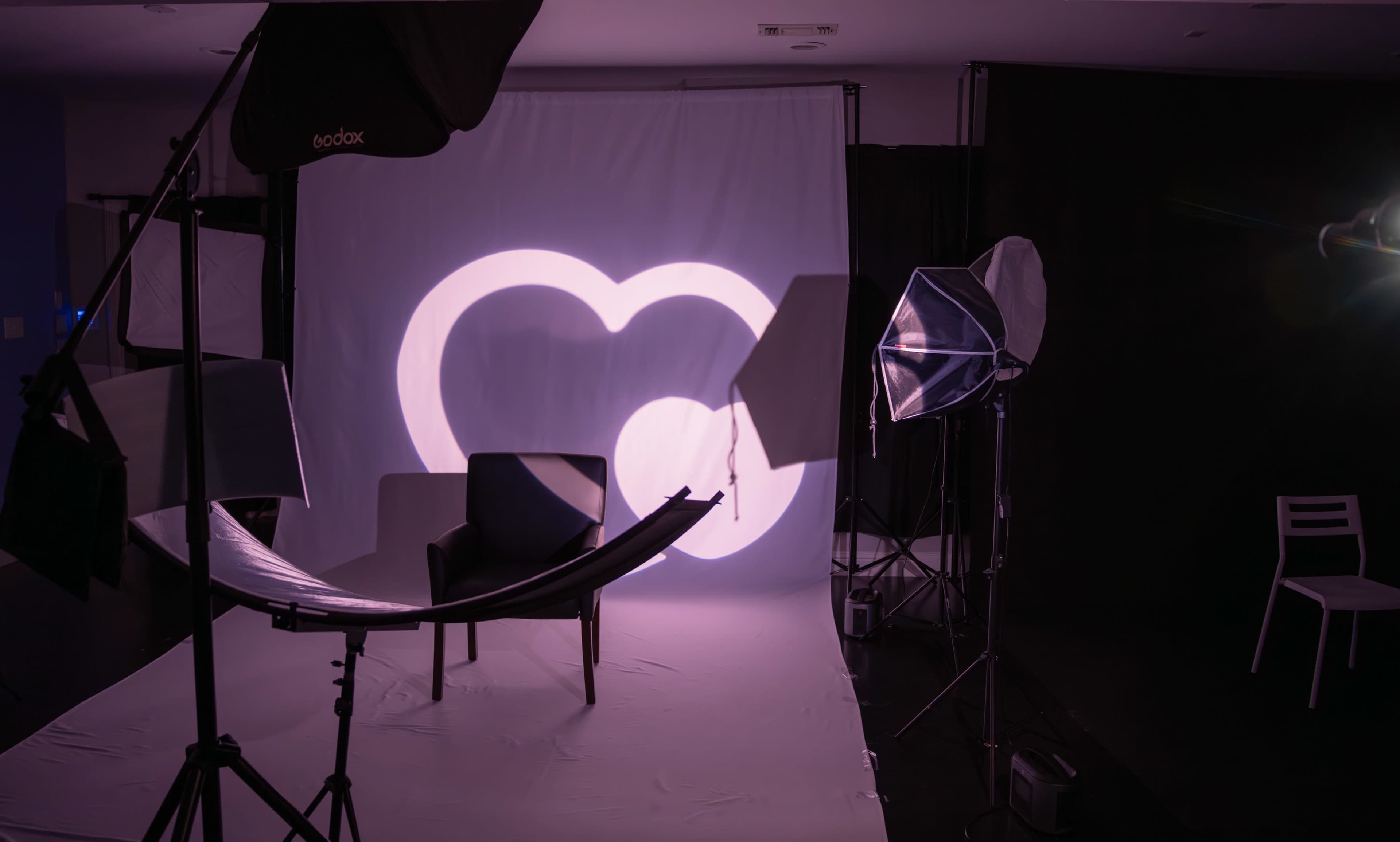A photography studio is set up with a white backdrop, a chair in the foreground, and various lighting equipment surrounding the scene.