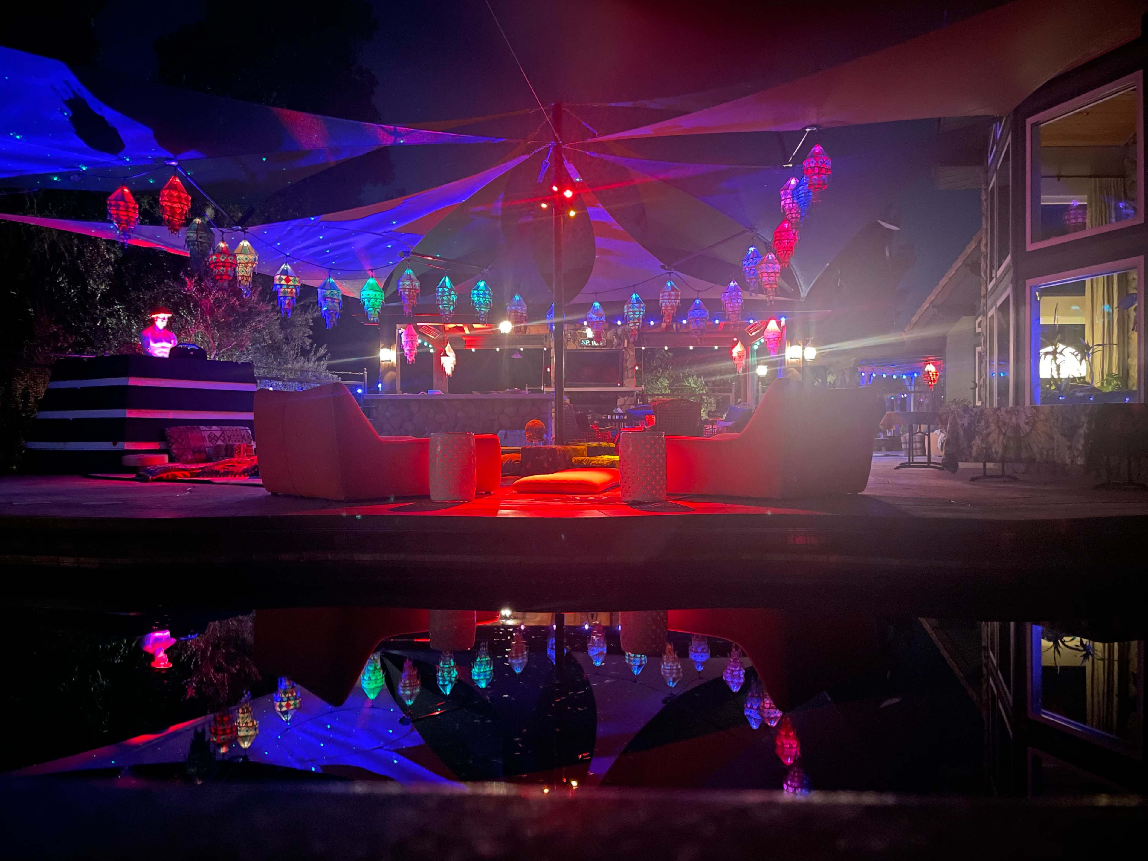 A nighttime patio scene illuminated by colorful lights, featuring two white chairs facing a pool that reflects the vibrant decorations overhead.