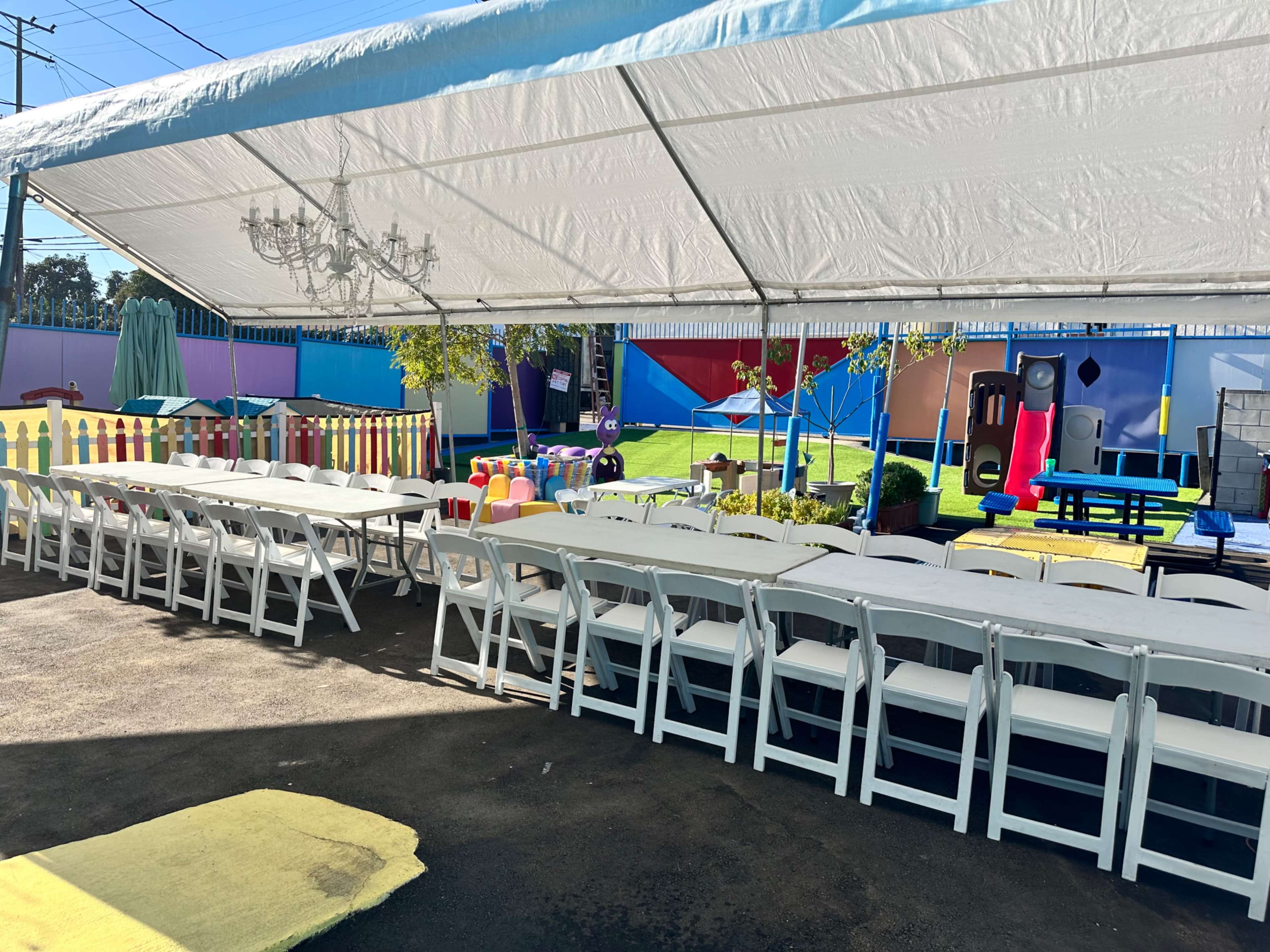 A large outdoor tent with several rows of white tables and chairs is set up next to a colorful play area featuring playground equipment and decorations.
