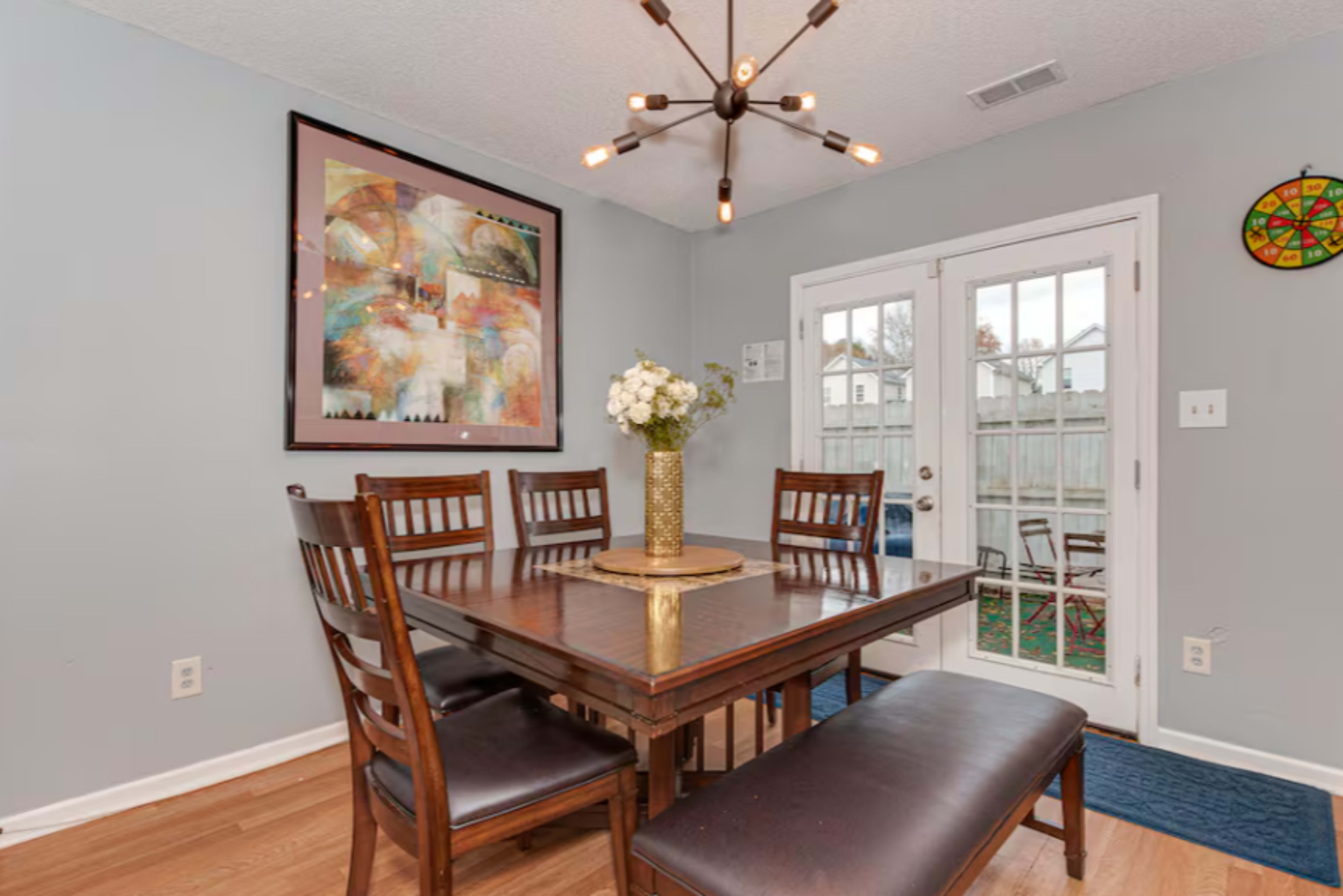 A dining area features a rectangular wooden table surrounded by six chairs, with a decorative light fixture above and a floral centerpiece on the table.