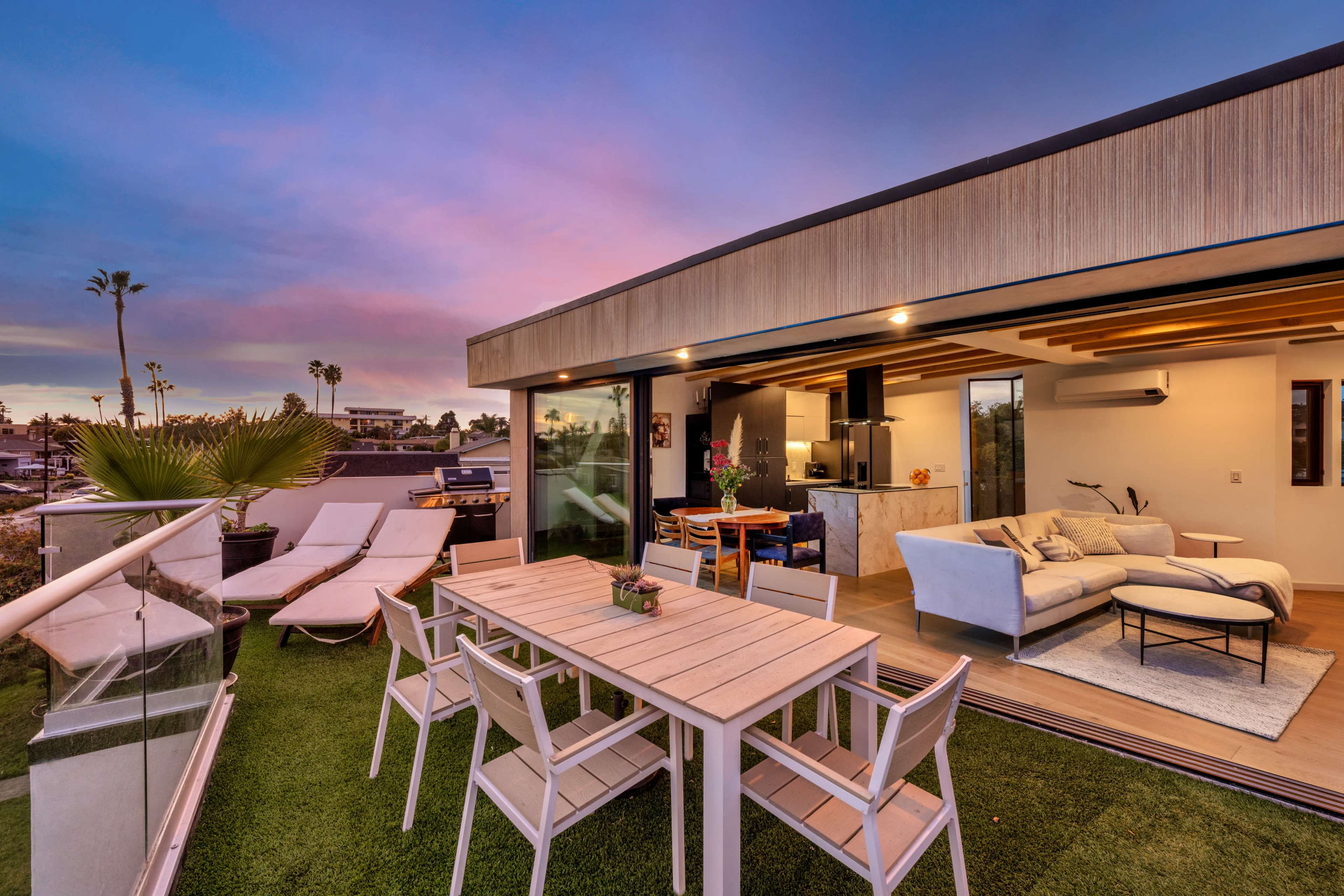 A modern rooftop patio features a dining table, lounge chairs, and a view of the sunset skyline.