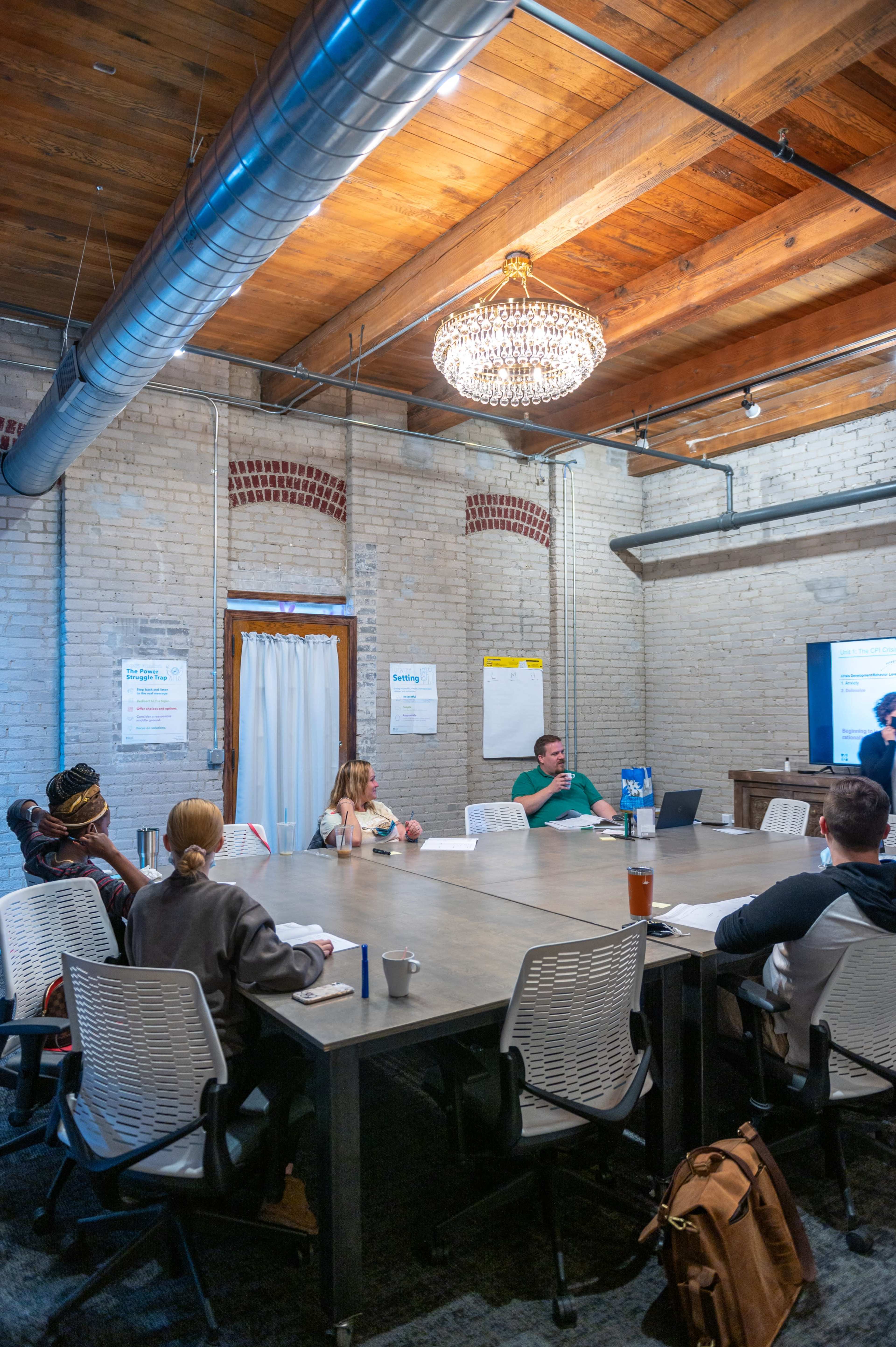 A group of people is seated around a large table in a conference room, with a presentation displayed on a screen at one end.