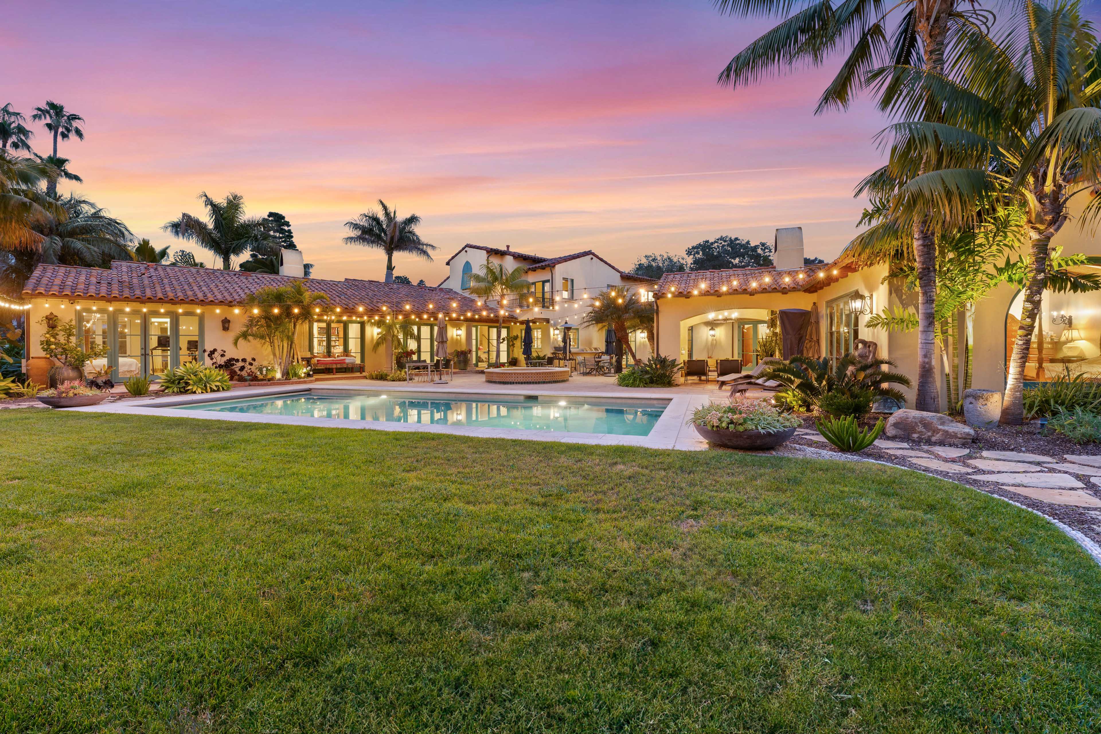 A spacious backyard features a swimming pool surrounded by lush grass, palm trees, and a well-lit patio area with a Mediterranean-style home in the background.