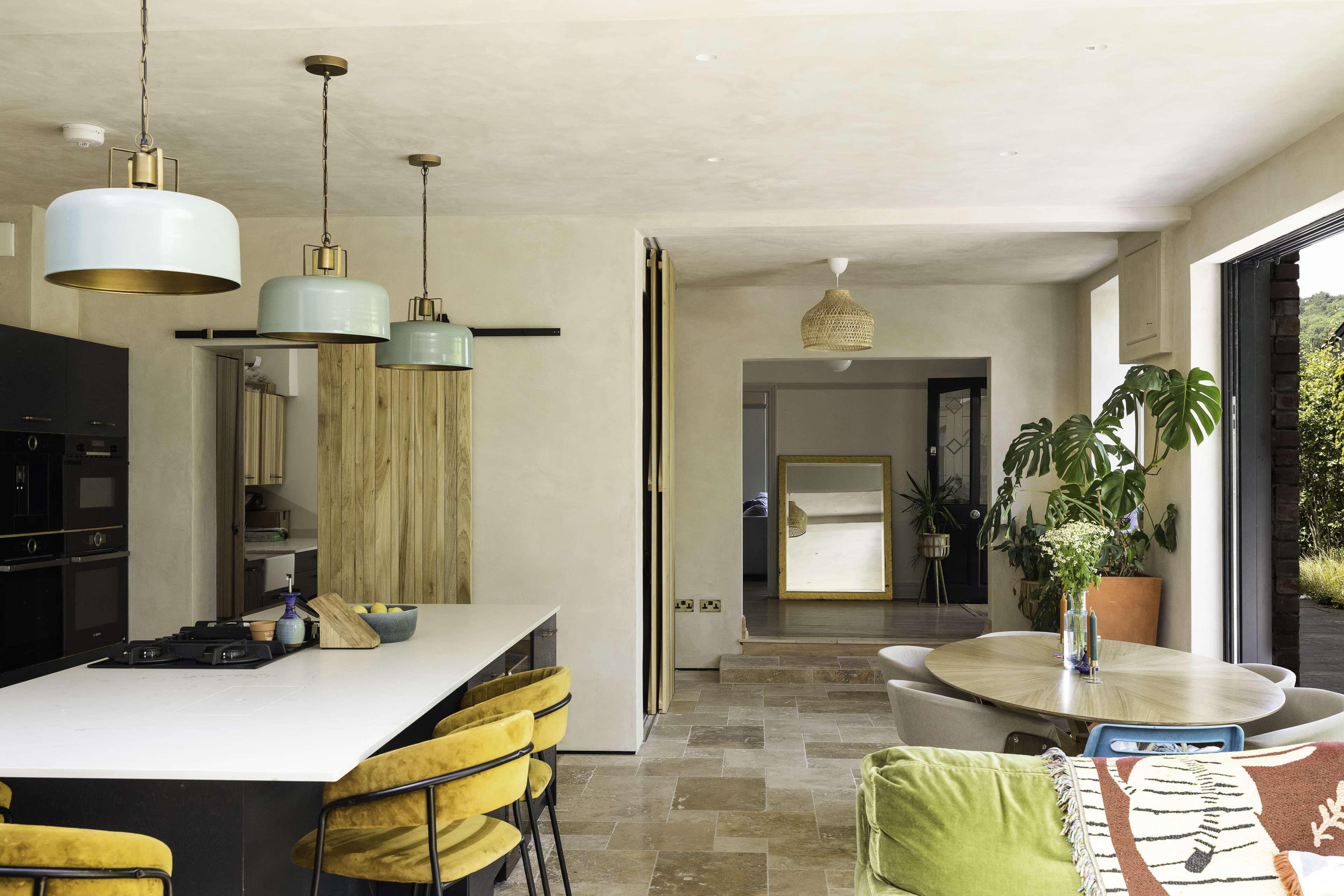 A modern kitchen and living area feature a central island with teal pendant lights, yellow upholstered chairs, and large glass doors opening to a garden.