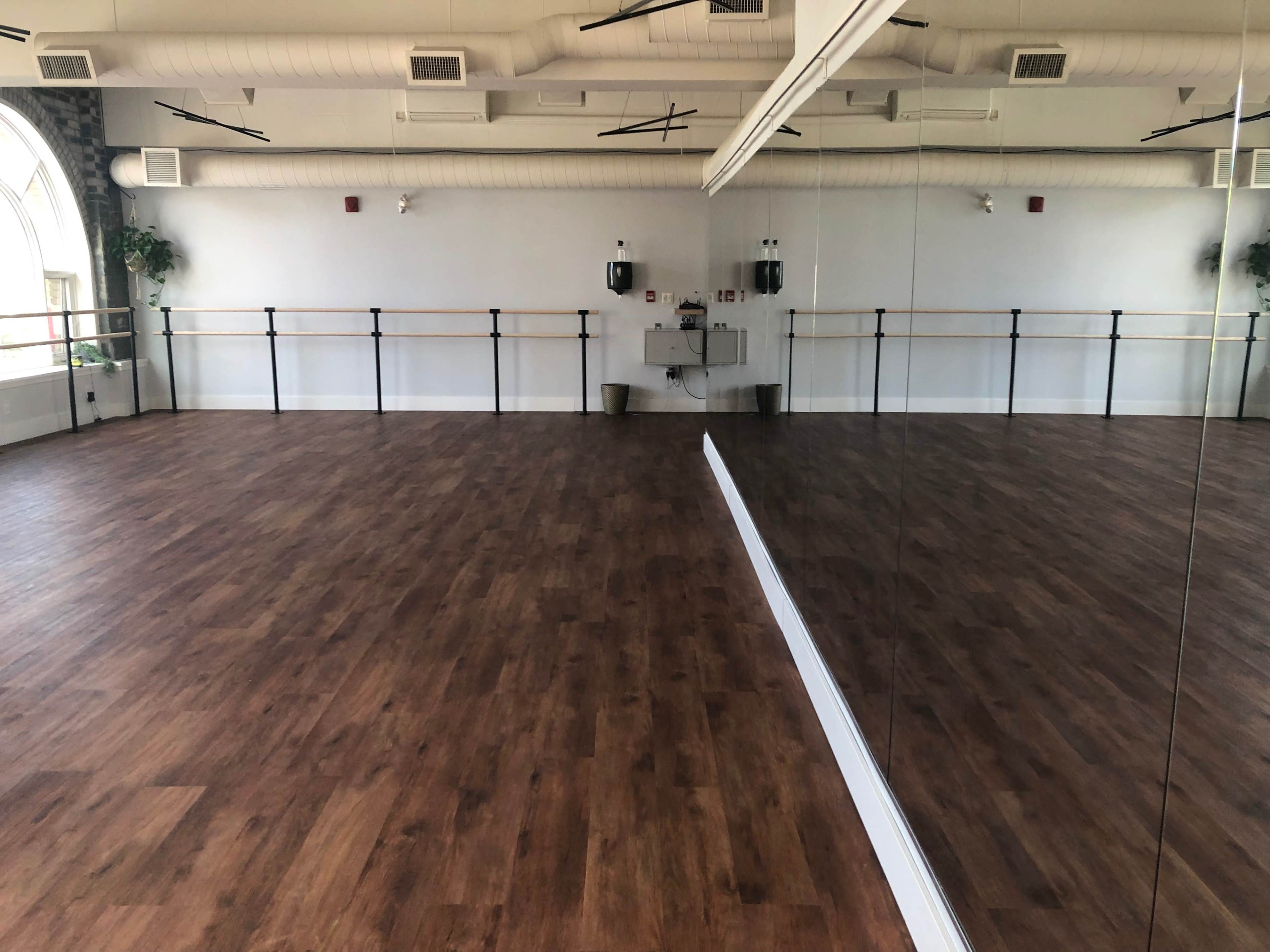 The image shows a spacious dance studio with wooden flooring, large mirrors on one wall, and ballet barres along the opposite wall.