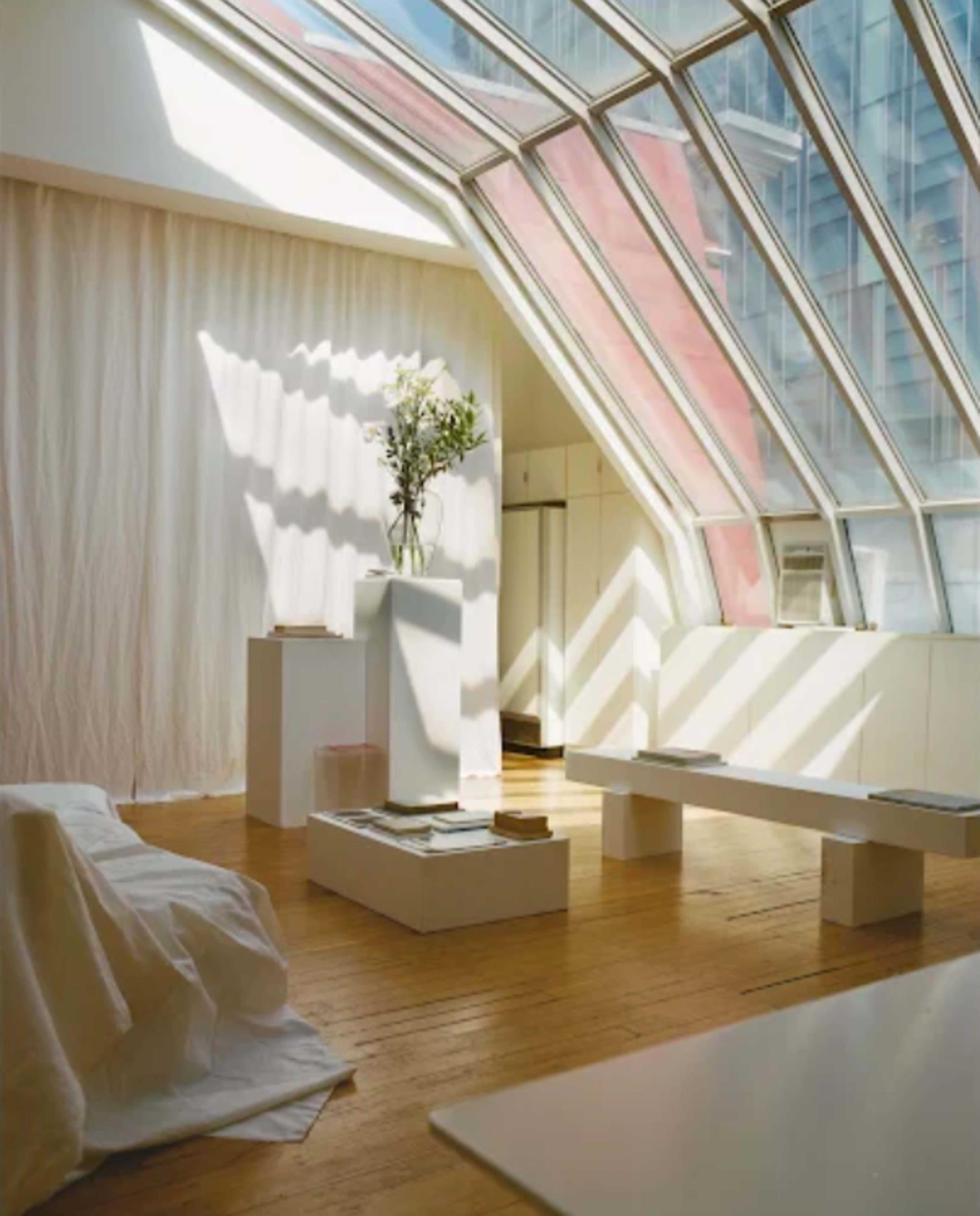 A bright room features a large glass skylight, white walls, a wooden floor, and minimalist furniture, including a bench and a display pedestal.