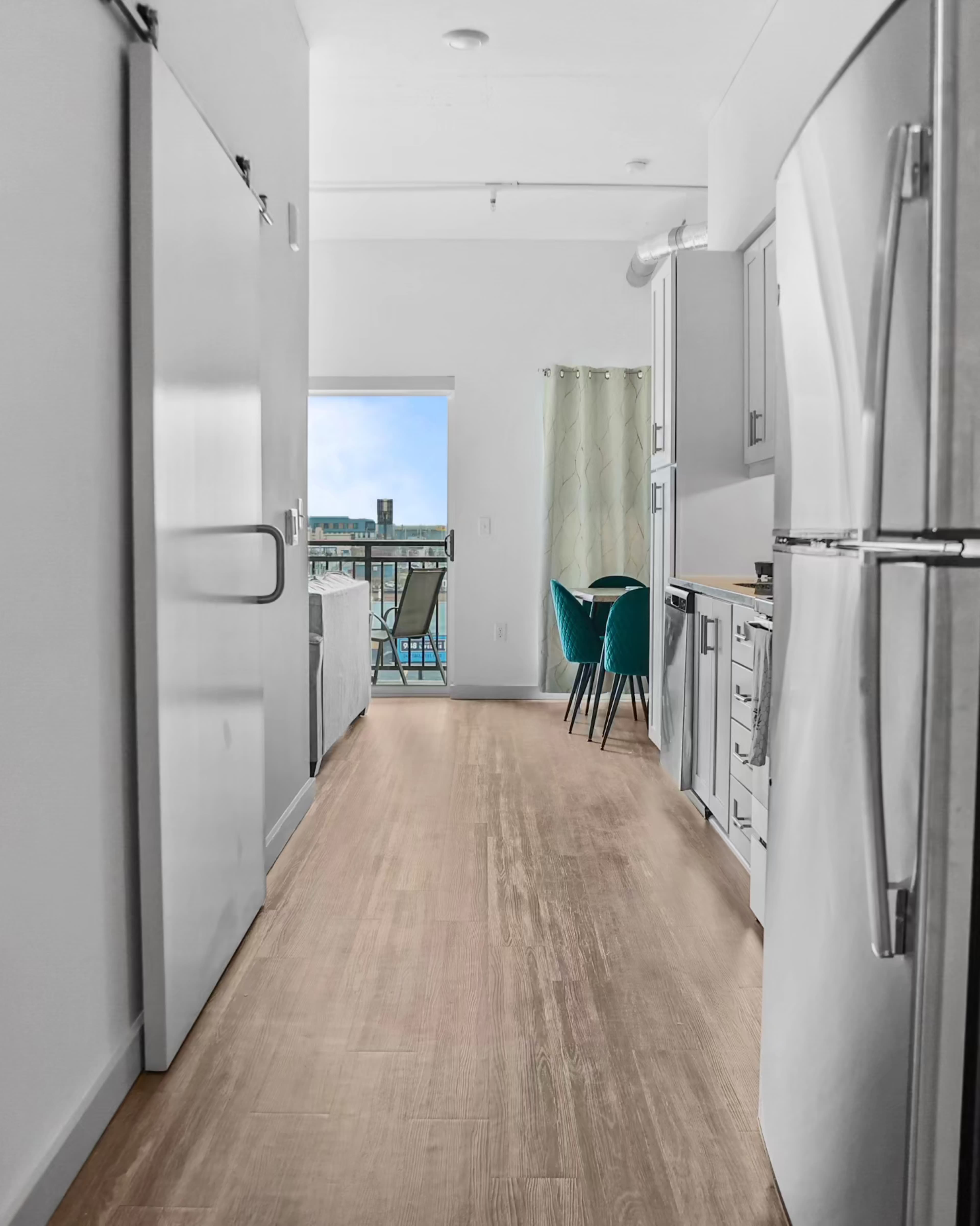 A narrow, bright hallway leads to a window with a view of the outdoors, flanked by a kitchen area on the right and a seating area on the left.