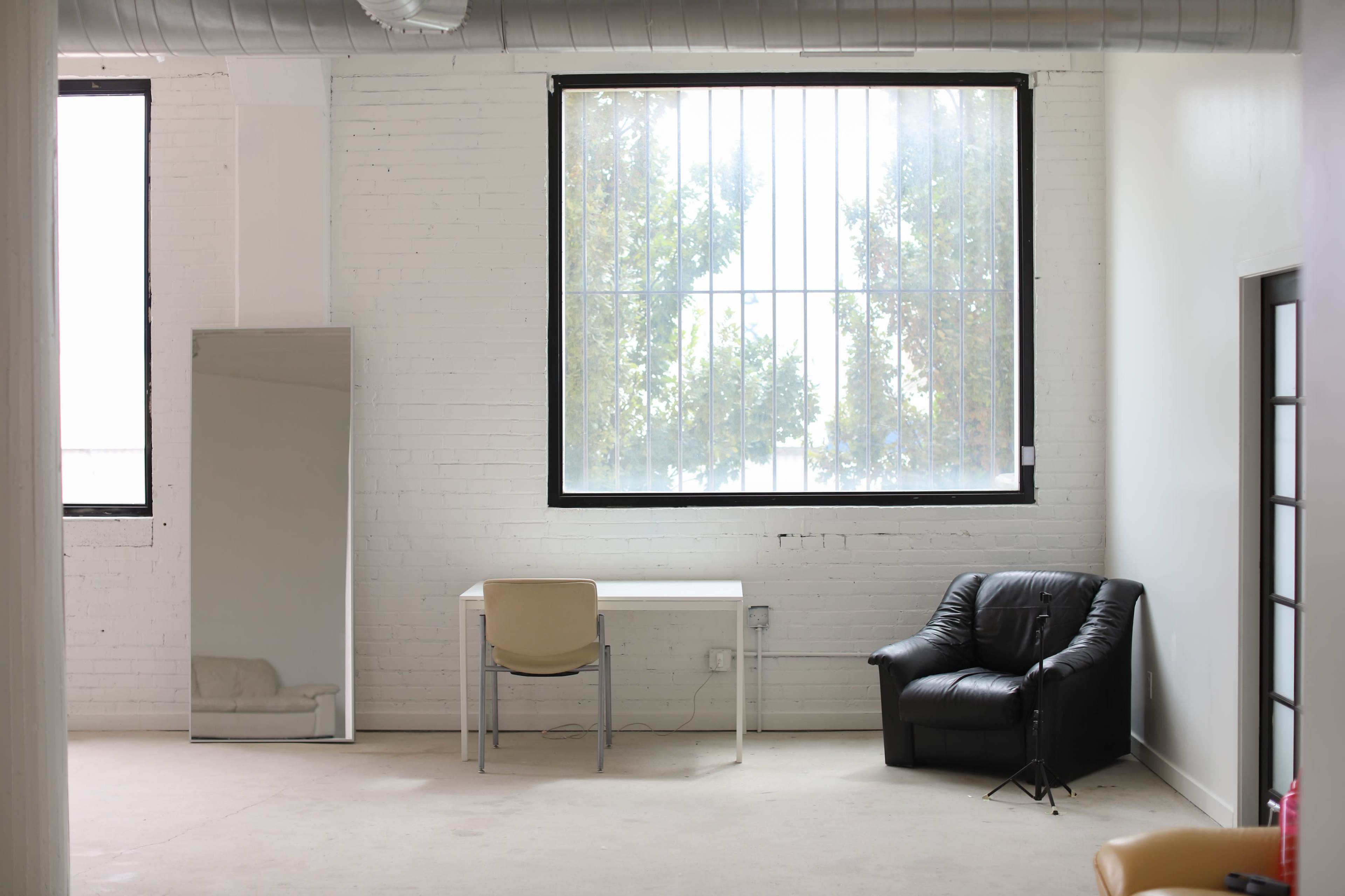A minimalistic room featuring a large window, a desk with a chair, a black leather armchair, and a full-length mirror.