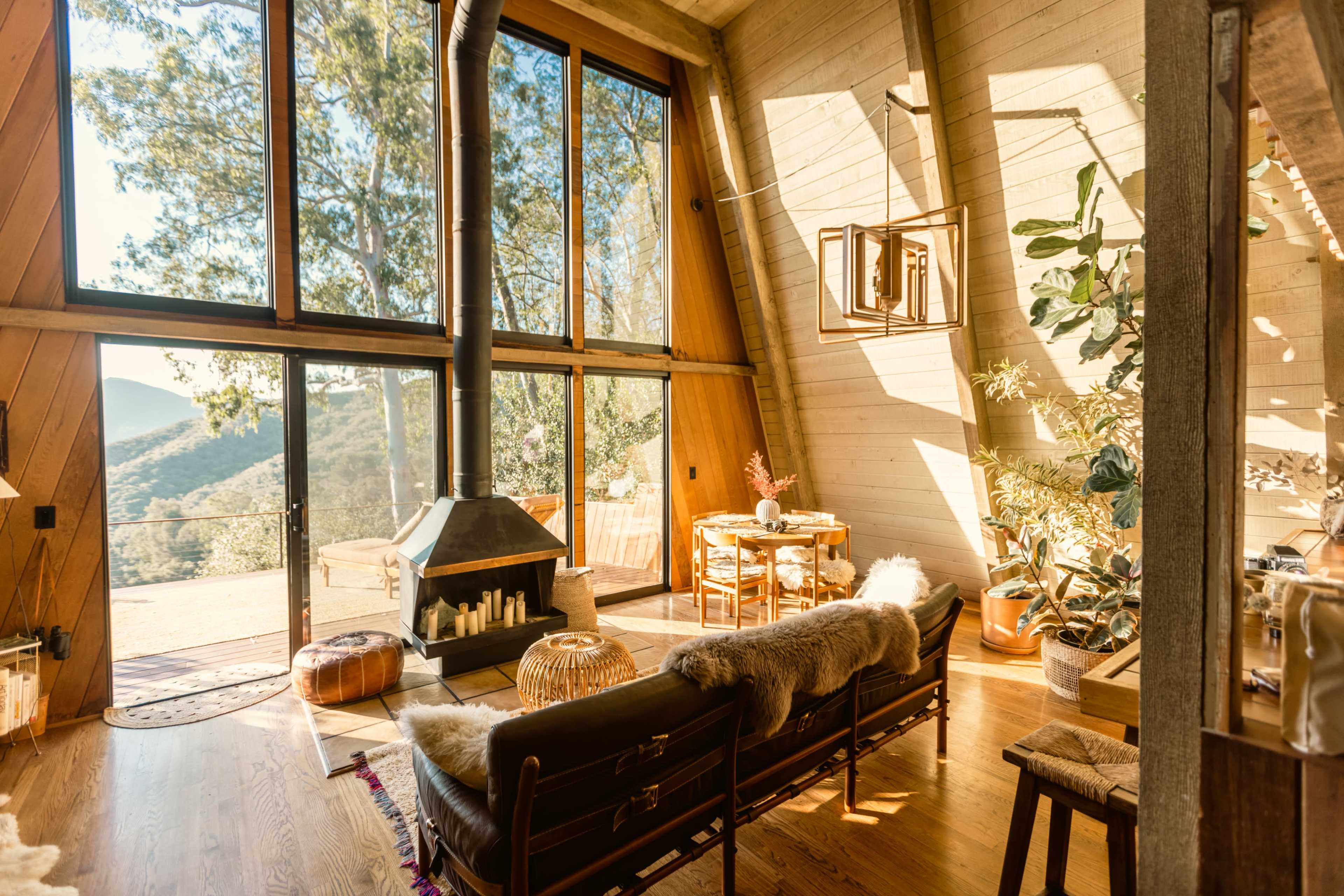 The interior of a modern cabin features large windows, a wood stove, and a cozy seating area with a wooden dining table.