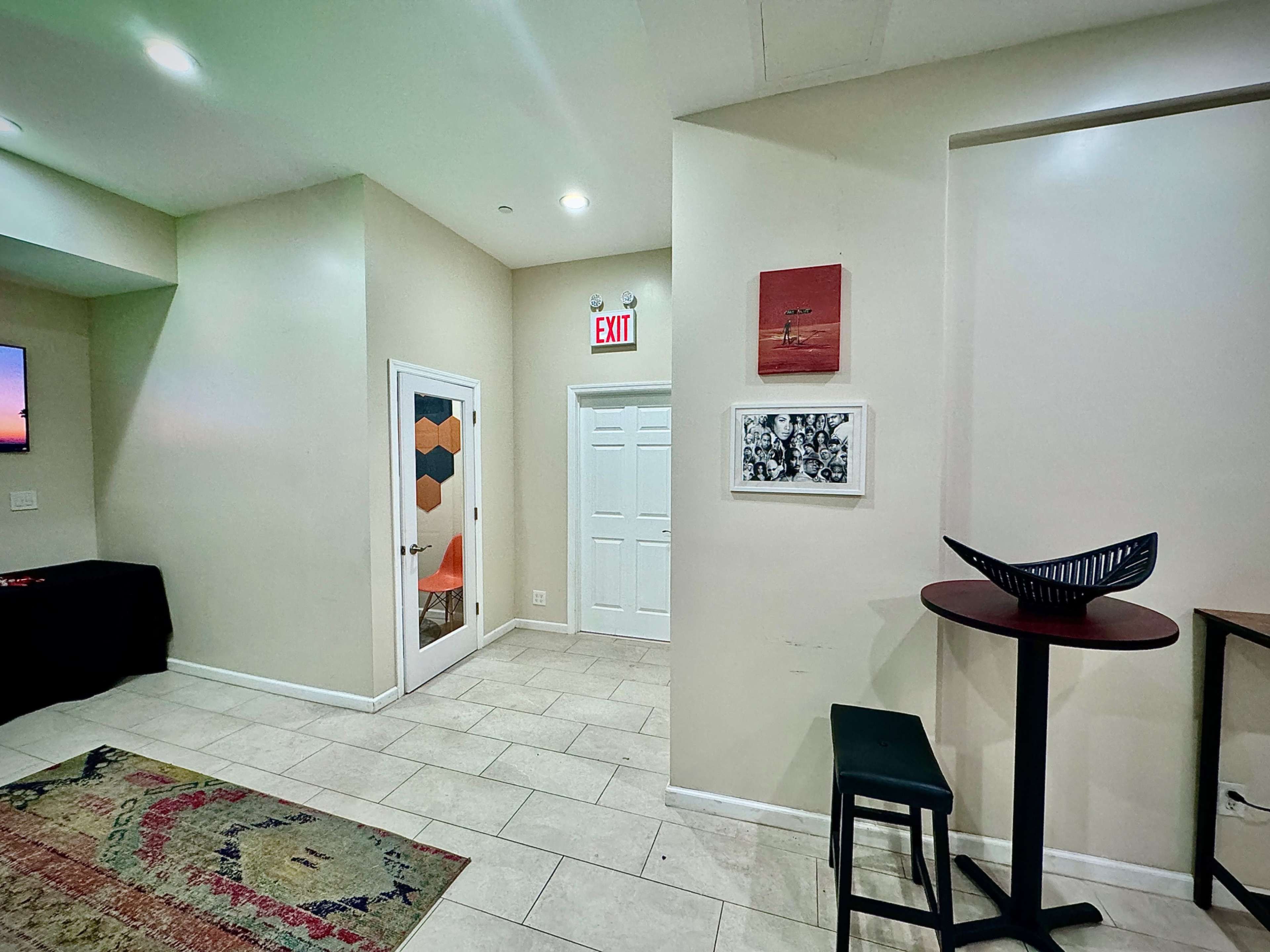 The image shows a well-lit interior space featuring a doorway, a wall with framed artwork, a small table with a black bowl, and a patterned rug on the tiled floor.