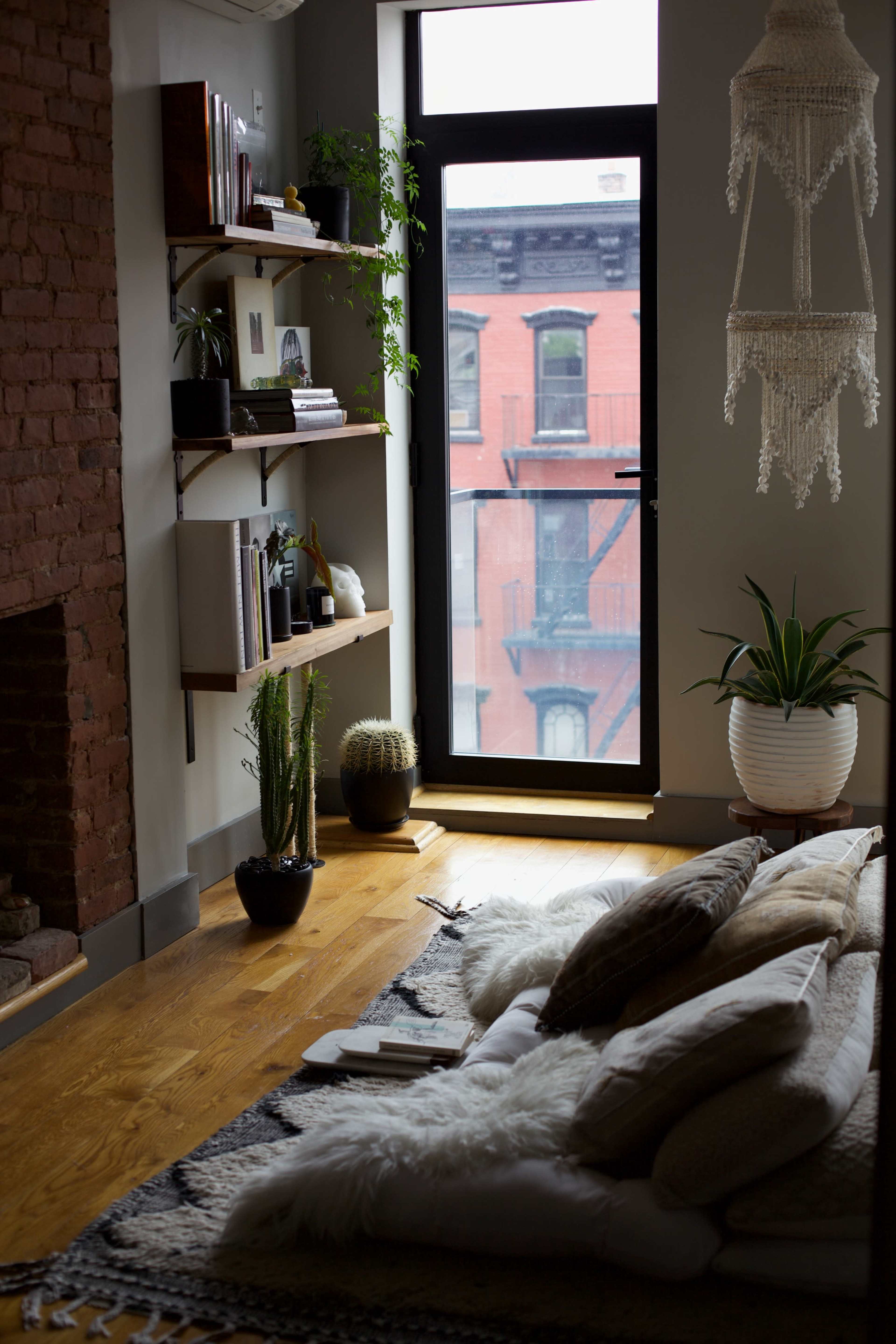A cozy living space features a large window with a view of a red brick building, a plush floor arrangement, and shelves displaying books and plants.