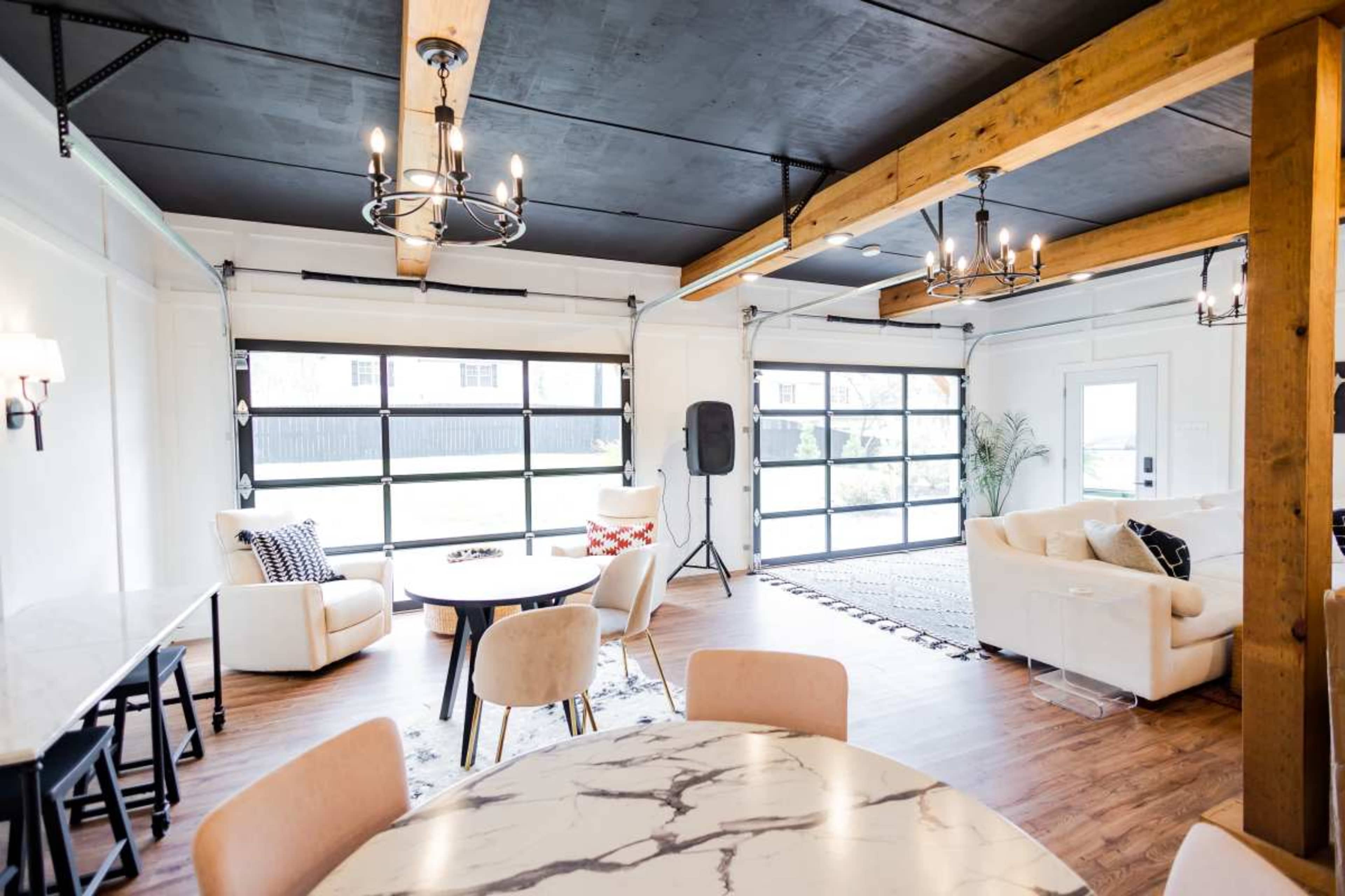 The image shows a spacious, modern living area with large garage doors, wooden beams, a round dining table, and comfortable seating arrangements.