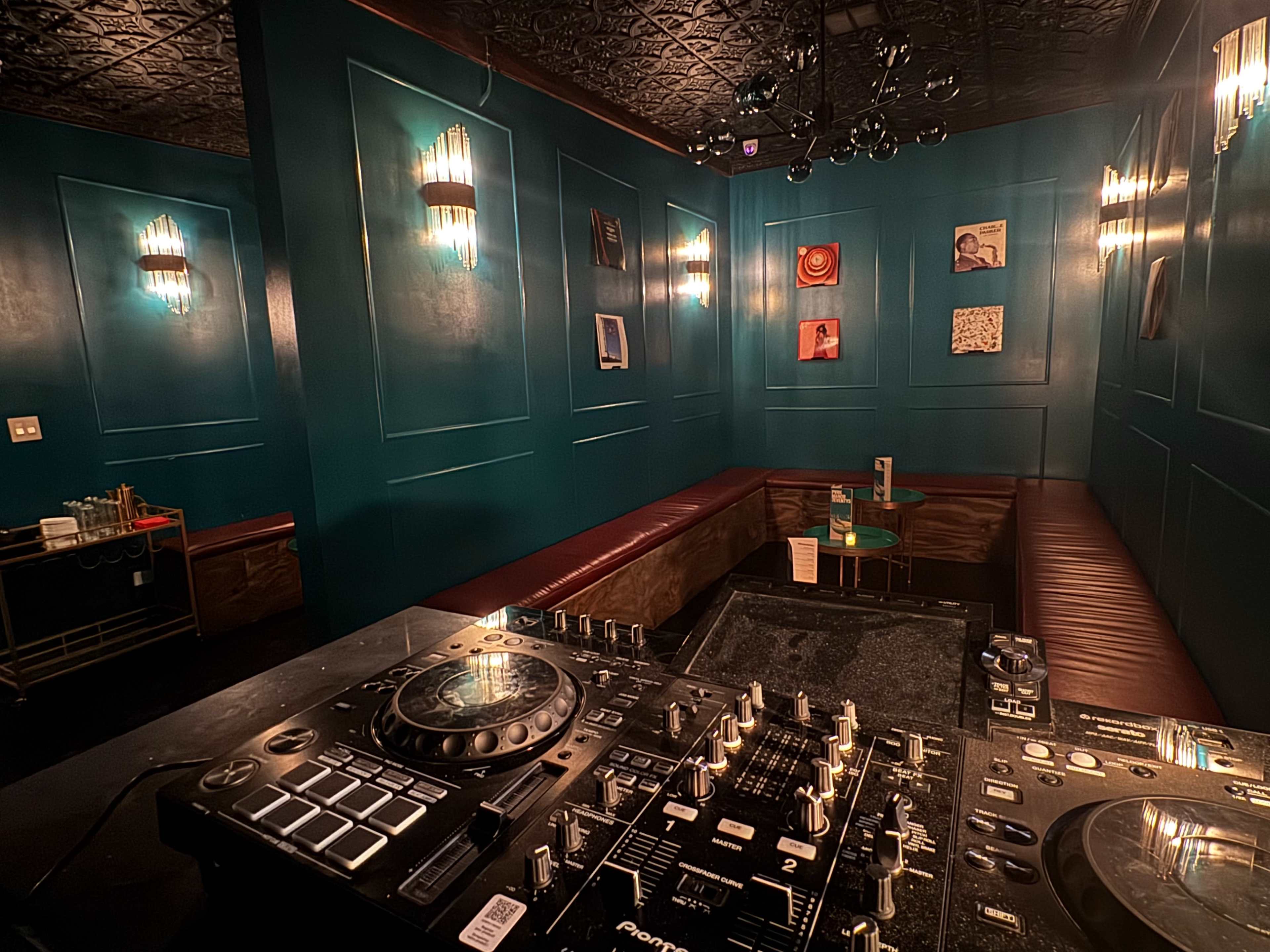 A DJ setup is positioned in the foreground of a dimly lit lounge featuring teal walls and cozy seating.
