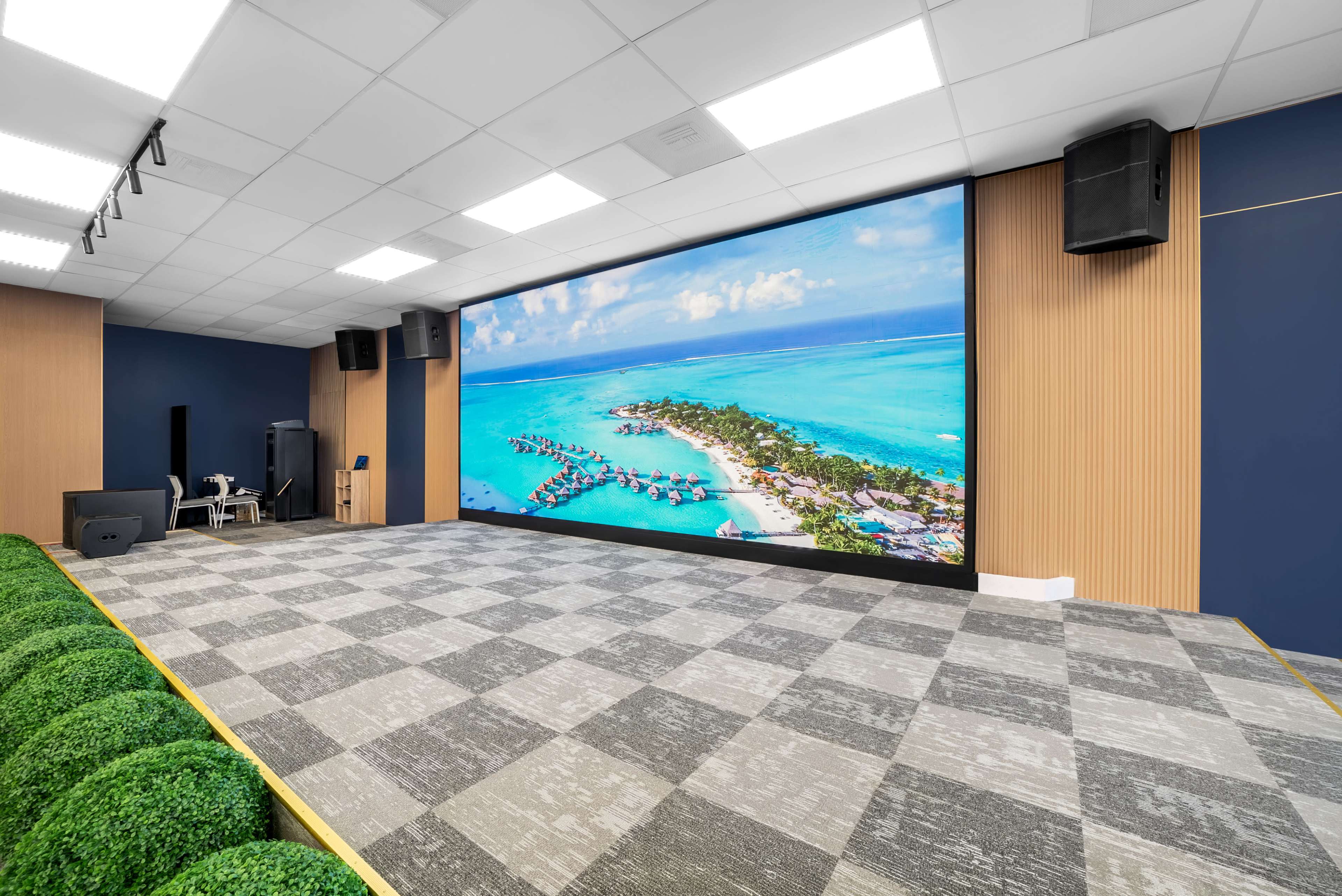 The image shows a modern interior space featuring a large screen displaying a tropical island view, with sound equipment on the walls and a patterned carpet below.