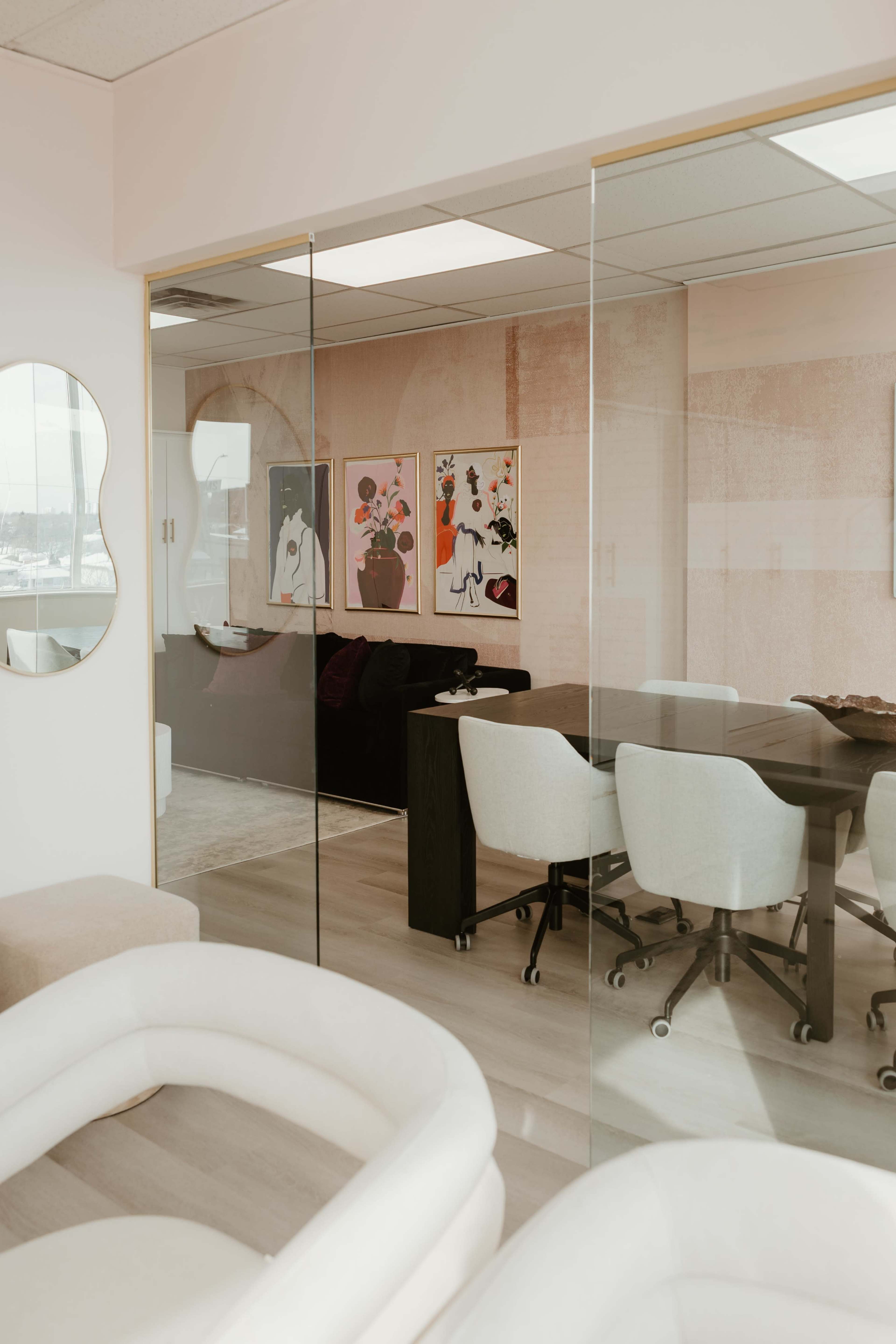 The image shows a modern office space with glass walls, featuring a conference table surrounded by chairs and two colorful framed artworks on the wall.