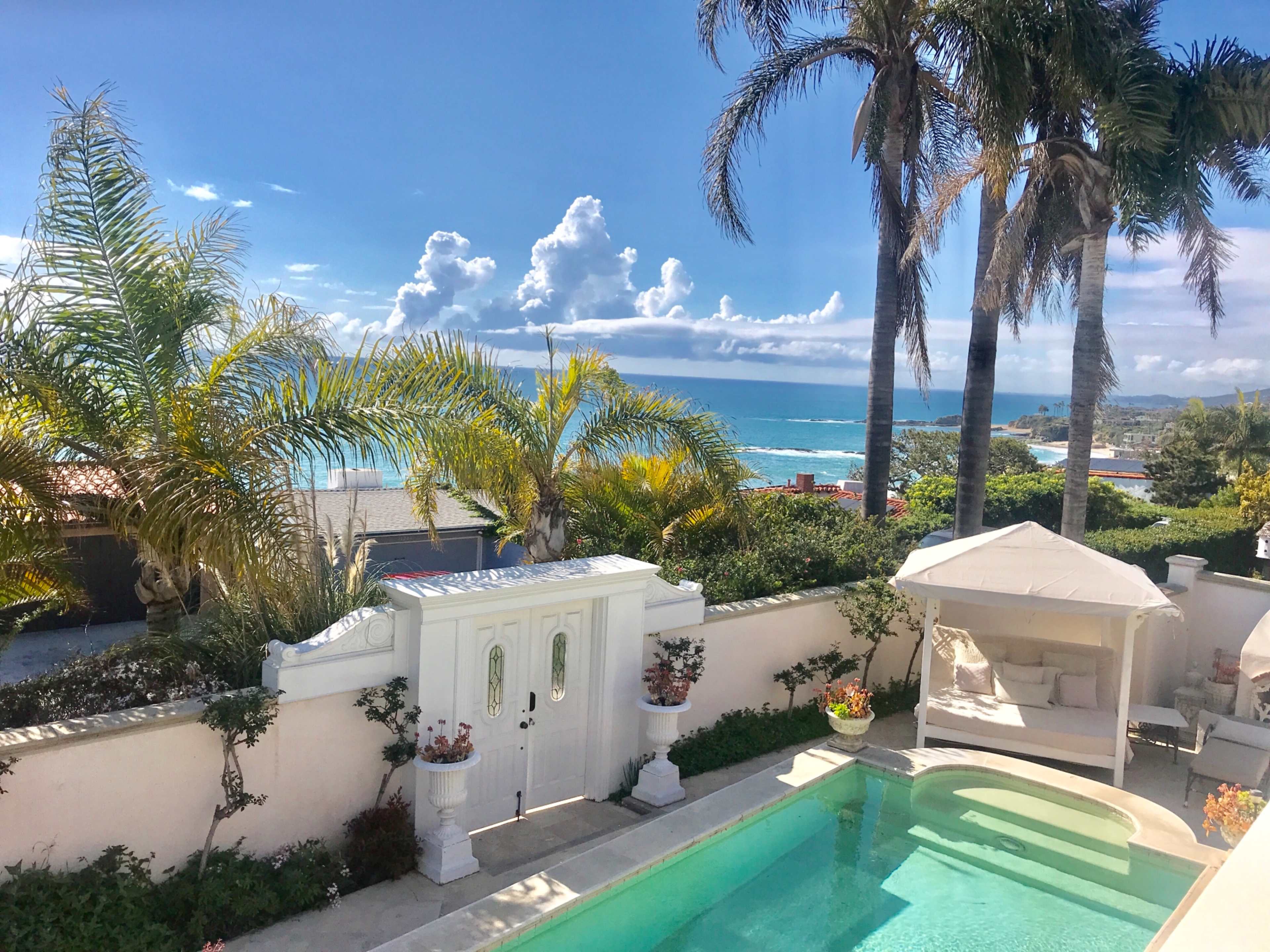 A view of a swimming pool surrounded by tropical plants, with the ocean and clouds visible in the background.