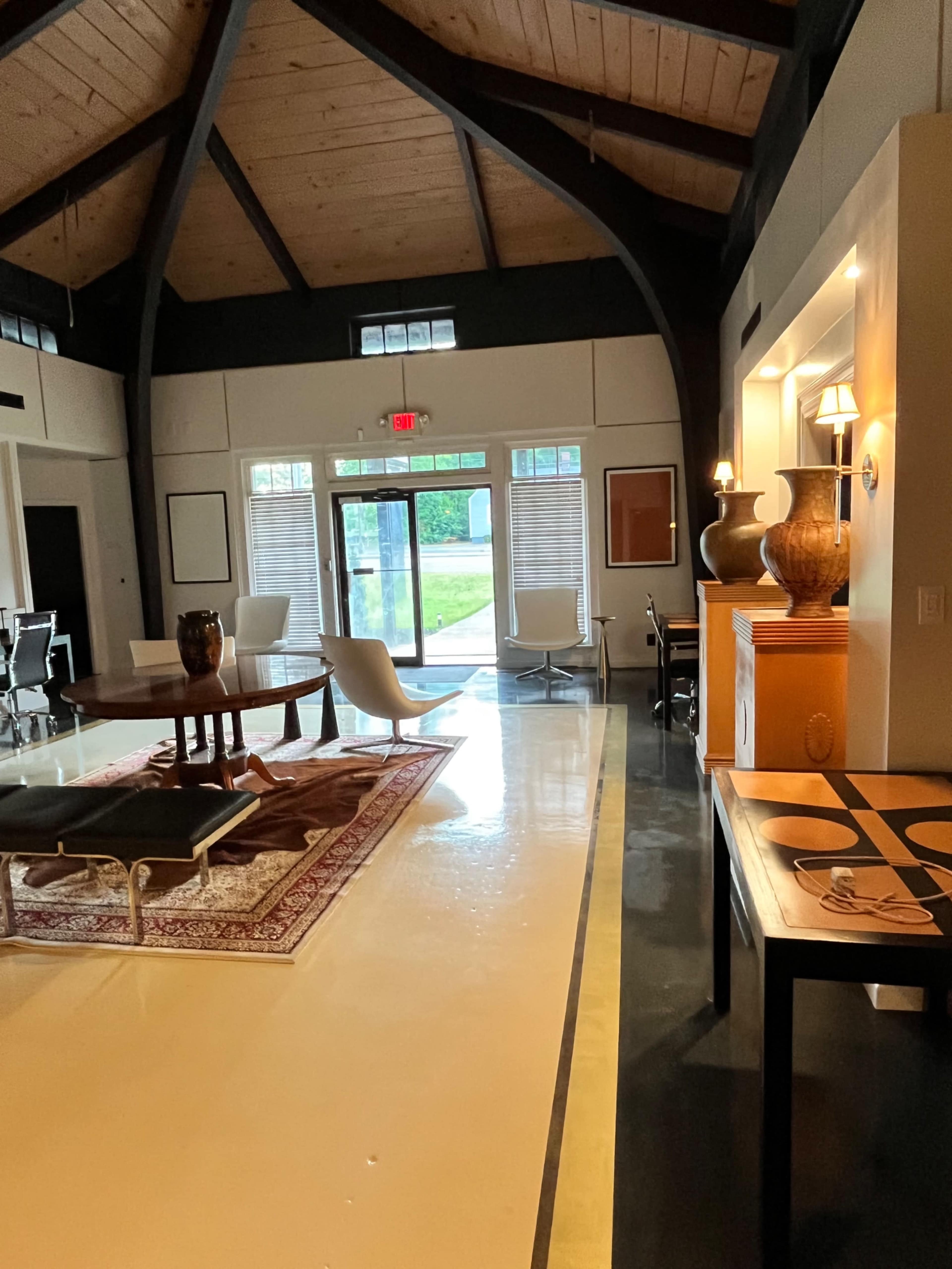 The image shows a spacious interior with high wooden ceilings, featuring a combination of modern furniture and decorative elements, and large windows allowing natural light to enter.