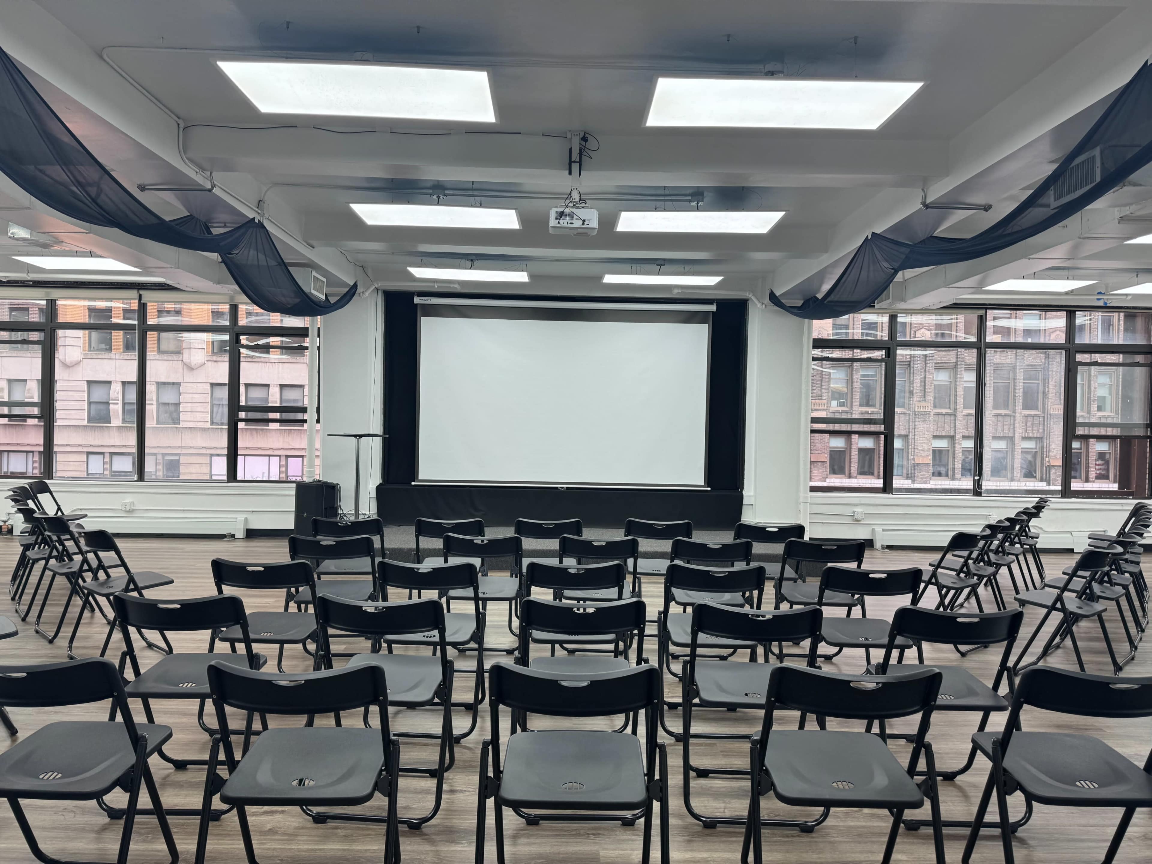 The image shows a spacious, well-lit room arranged with black folding chairs facing a blank projection screen at the front.
