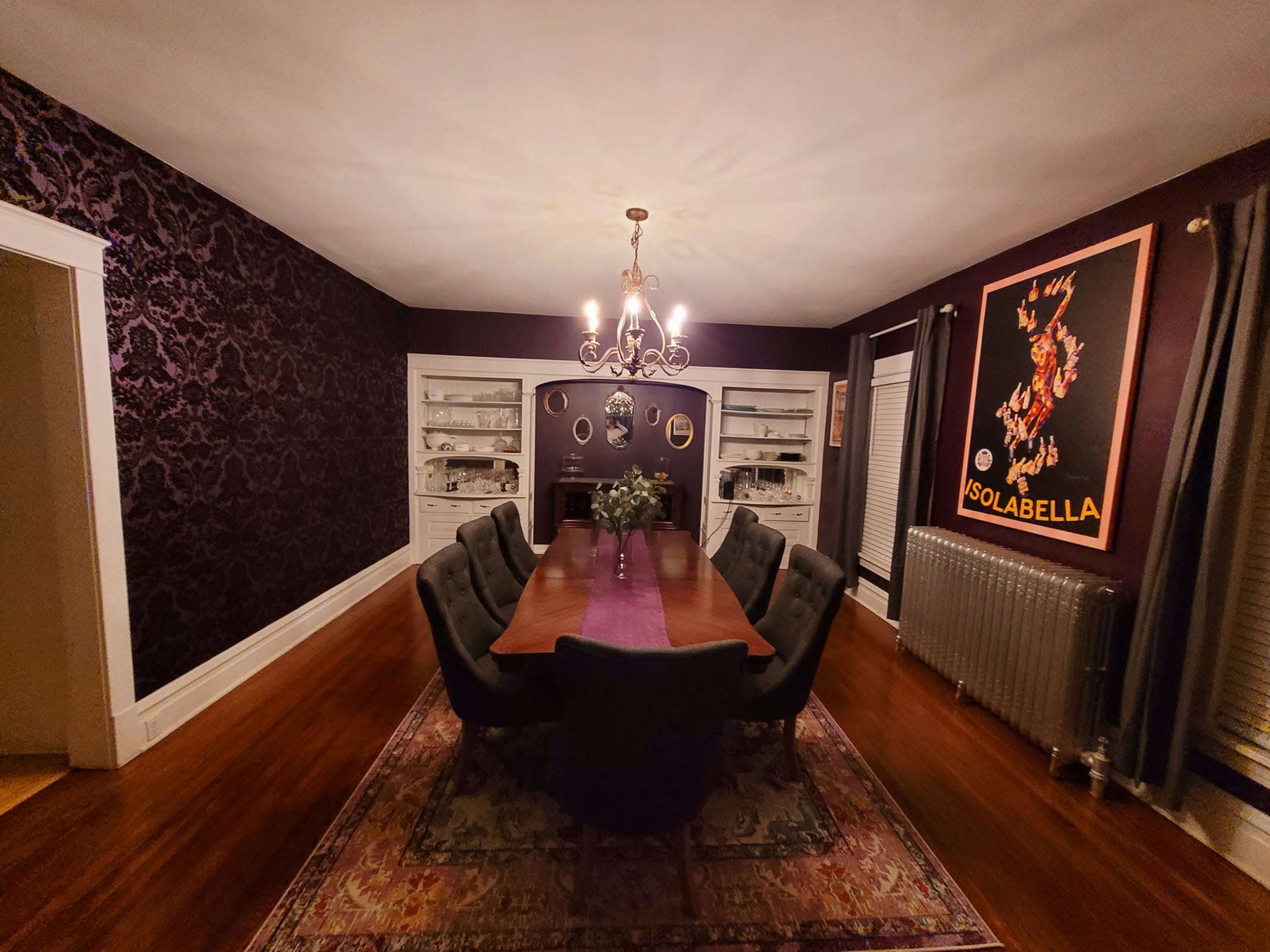 A dining room features a long wooden table surrounded by upholstered chairs, with dark patterned walls, a chandelier, and a vintage poster on display.