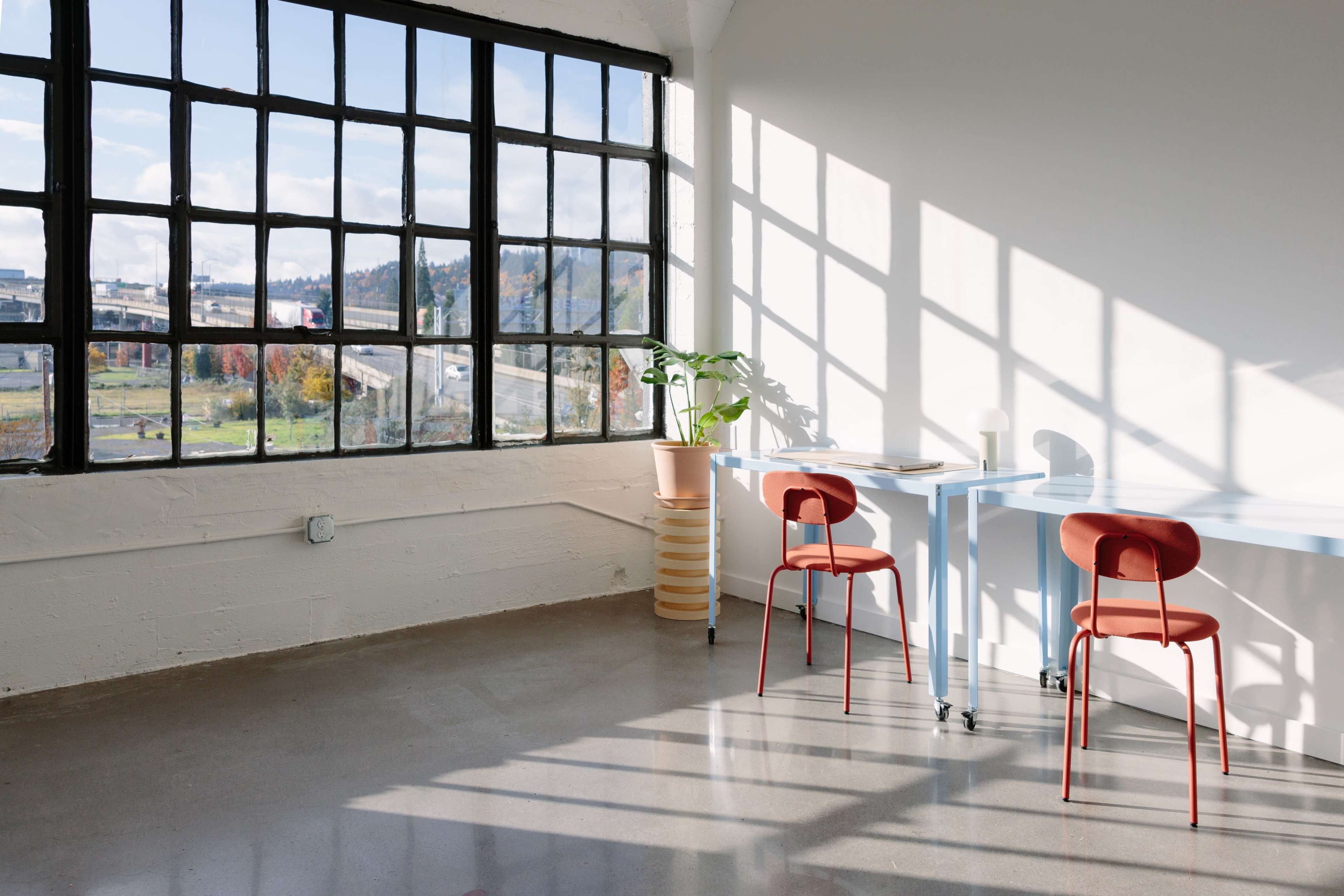 A brightly lit room features a large window with industrial frames, two blue tables, and red chairs, along with a potted plant beside the window.