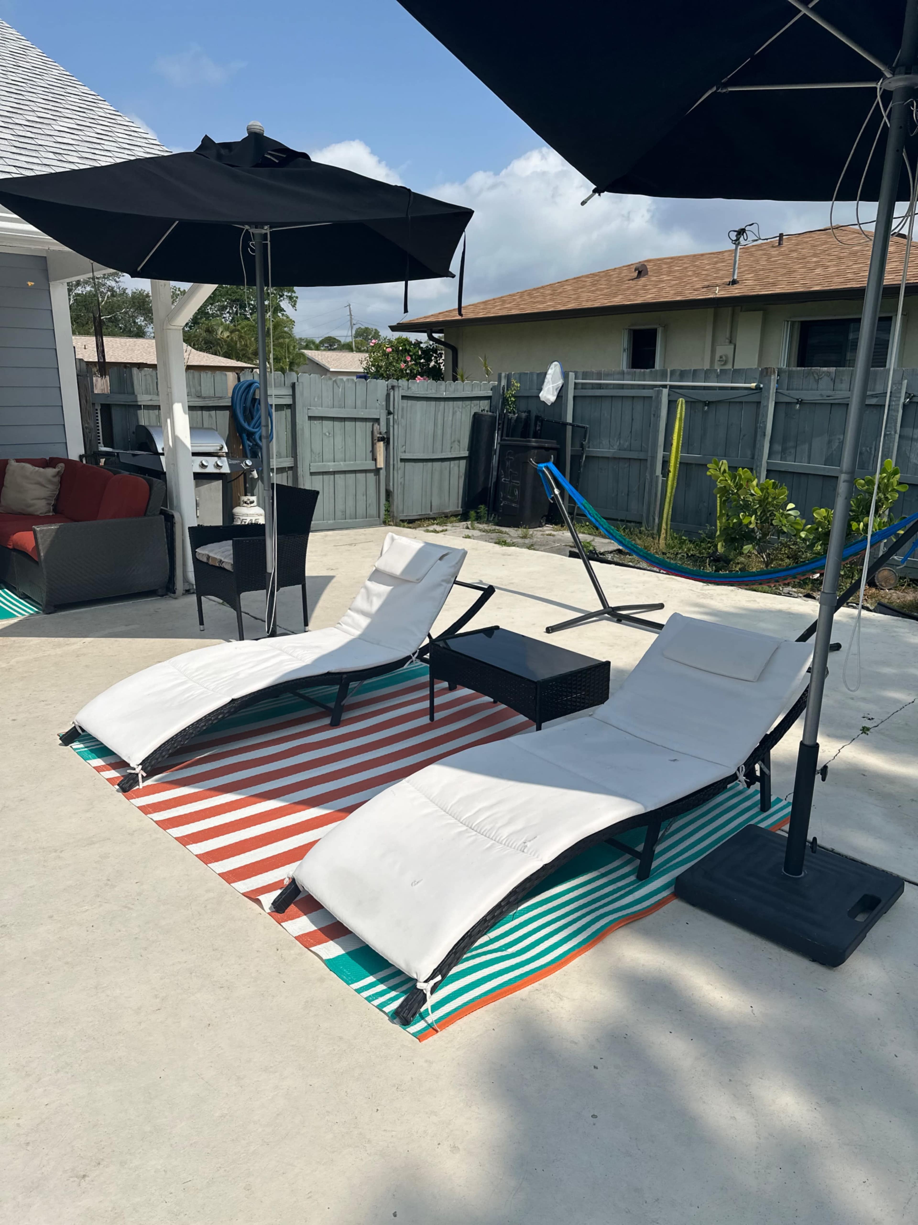 Two white lounge chairs positioned on a striped outdoor rug in a backyard with a shaded area provided by an umbrella.