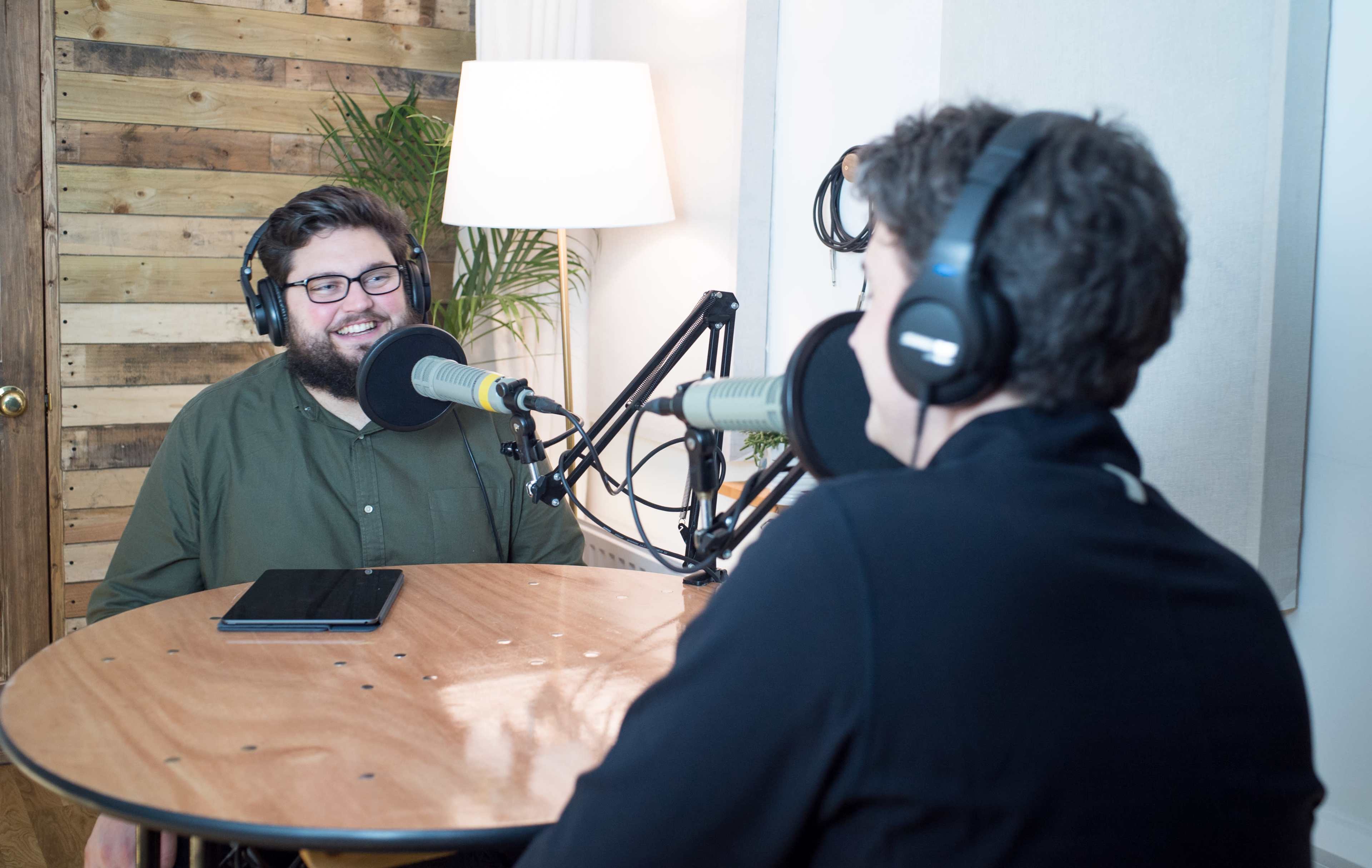 Two men are sitting at a round wooden table with microphones in front of them, engaged in a conversation in a podcast recording studio.
