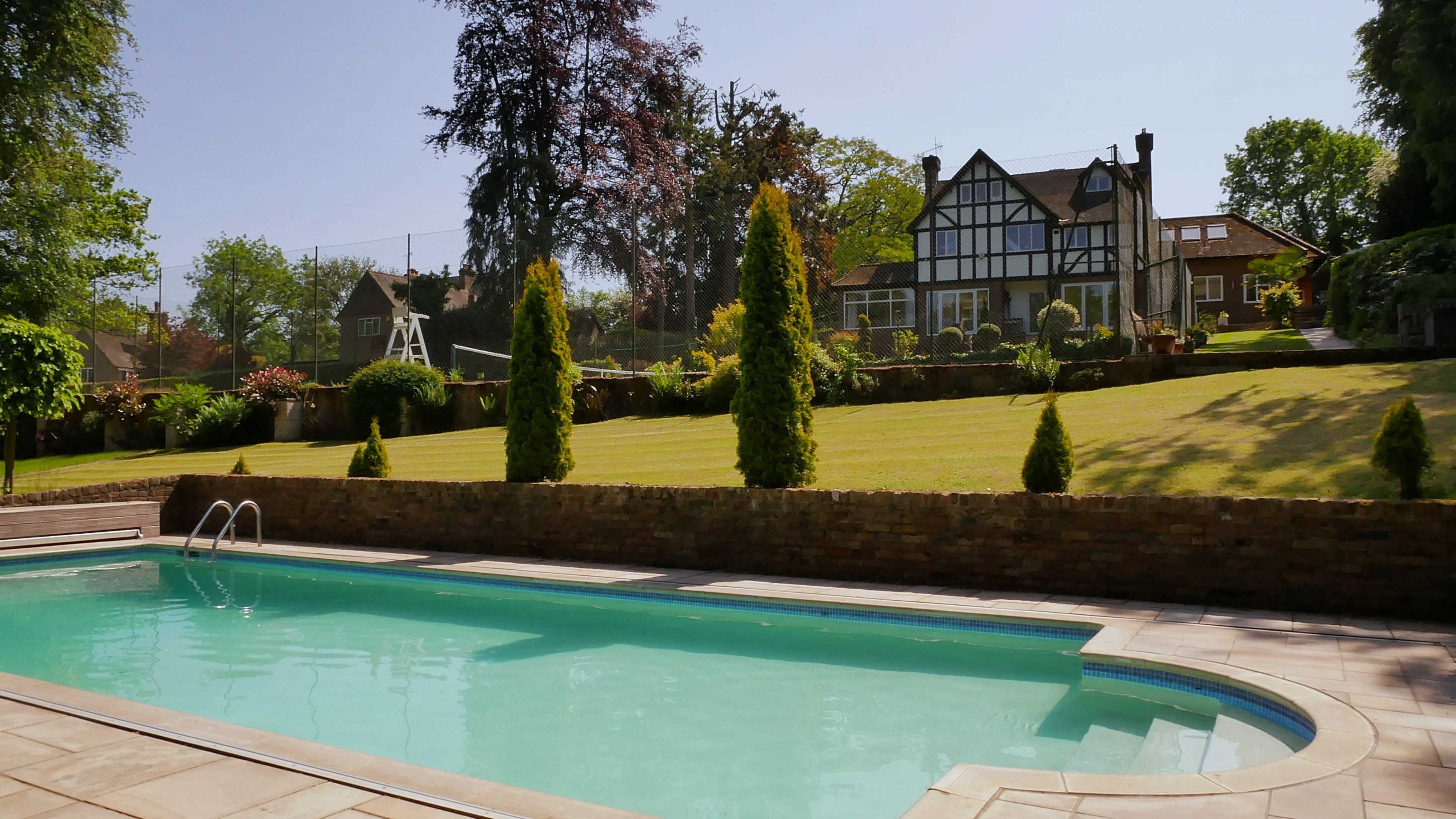 A clear swimming pool is situated in the foreground, with a landscaped garden and a large house in the background.