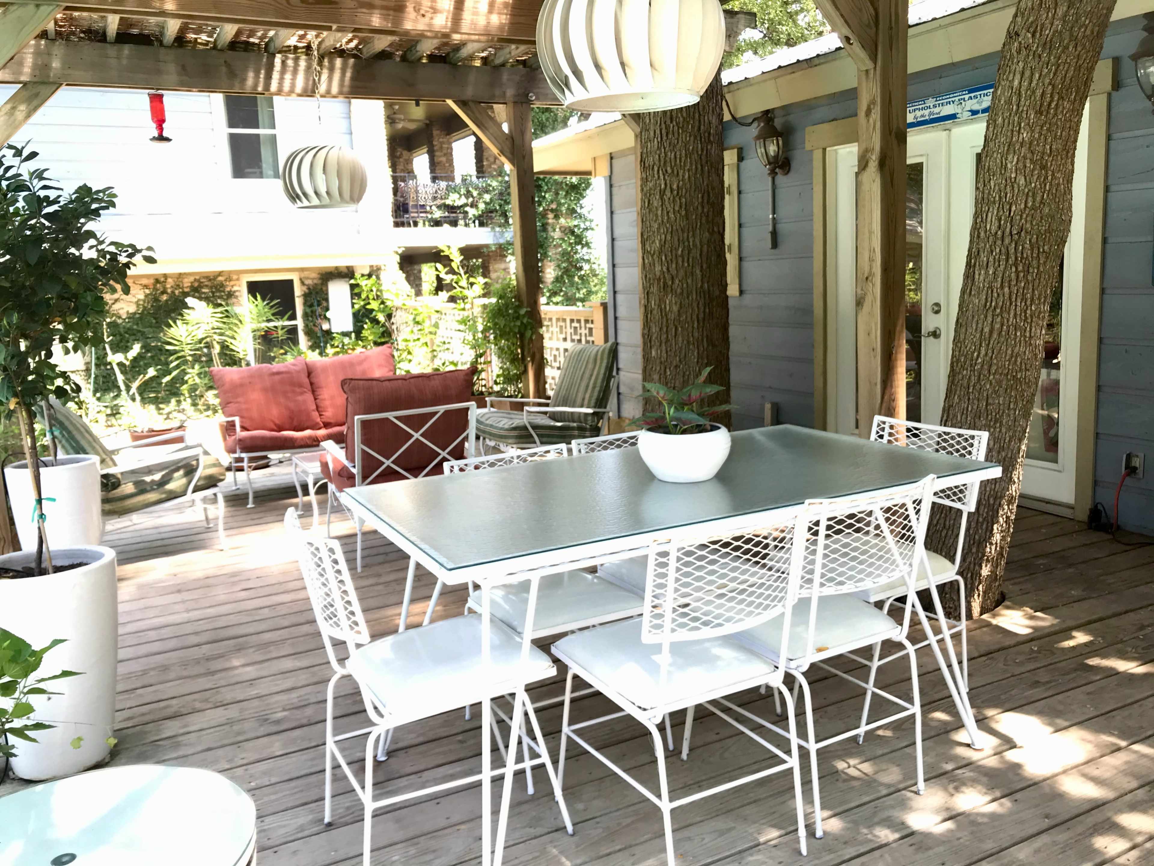 A patio area with a glass-top dining table surrounded by white metal chairs, complemented by potted plants and a seating arrangement nearby.