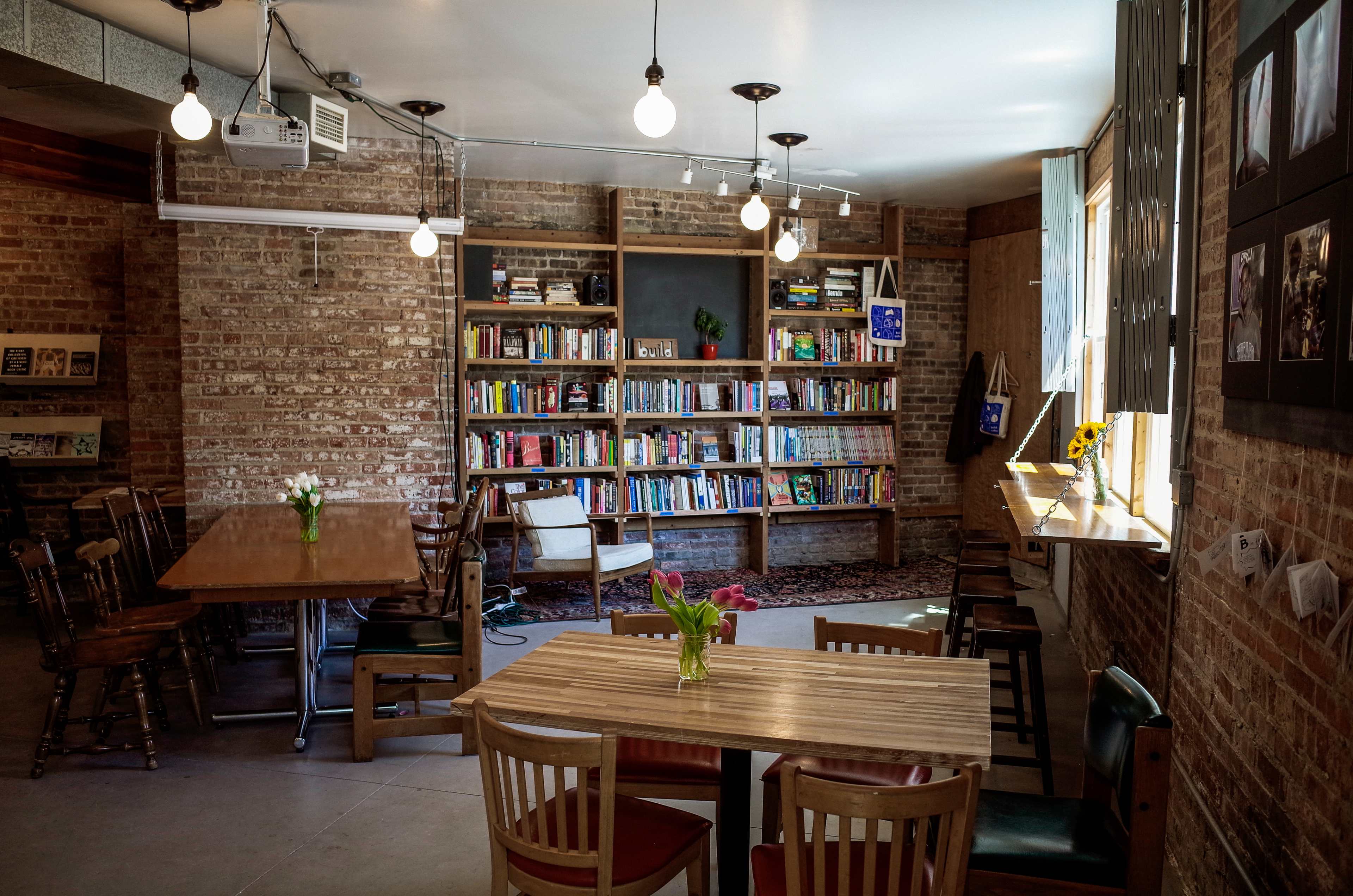 The scene features a cozy café with wooden tables and chairs, illuminated by hanging light bulbs, and a bookshelf filled with books against a brick wall.