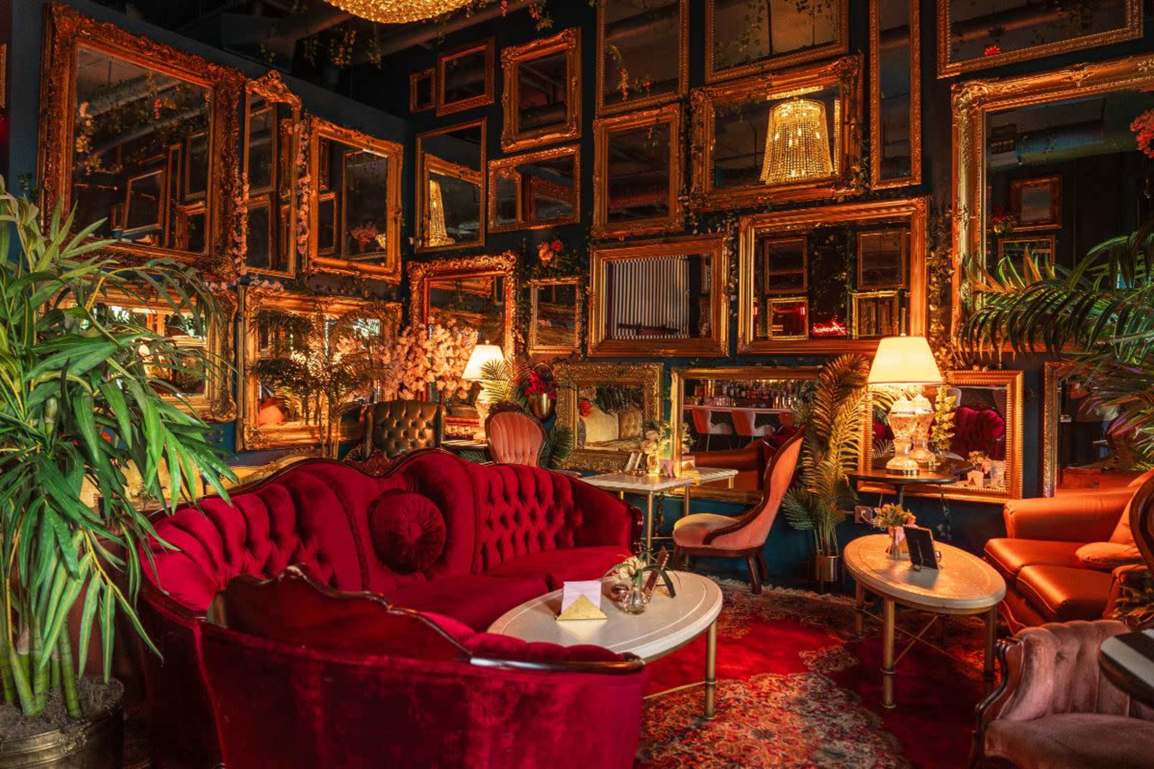 The image features a stylish interior with a plush red sofa, ornate mirrors lining the walls, and warm lighting from lamps and chandeliers.