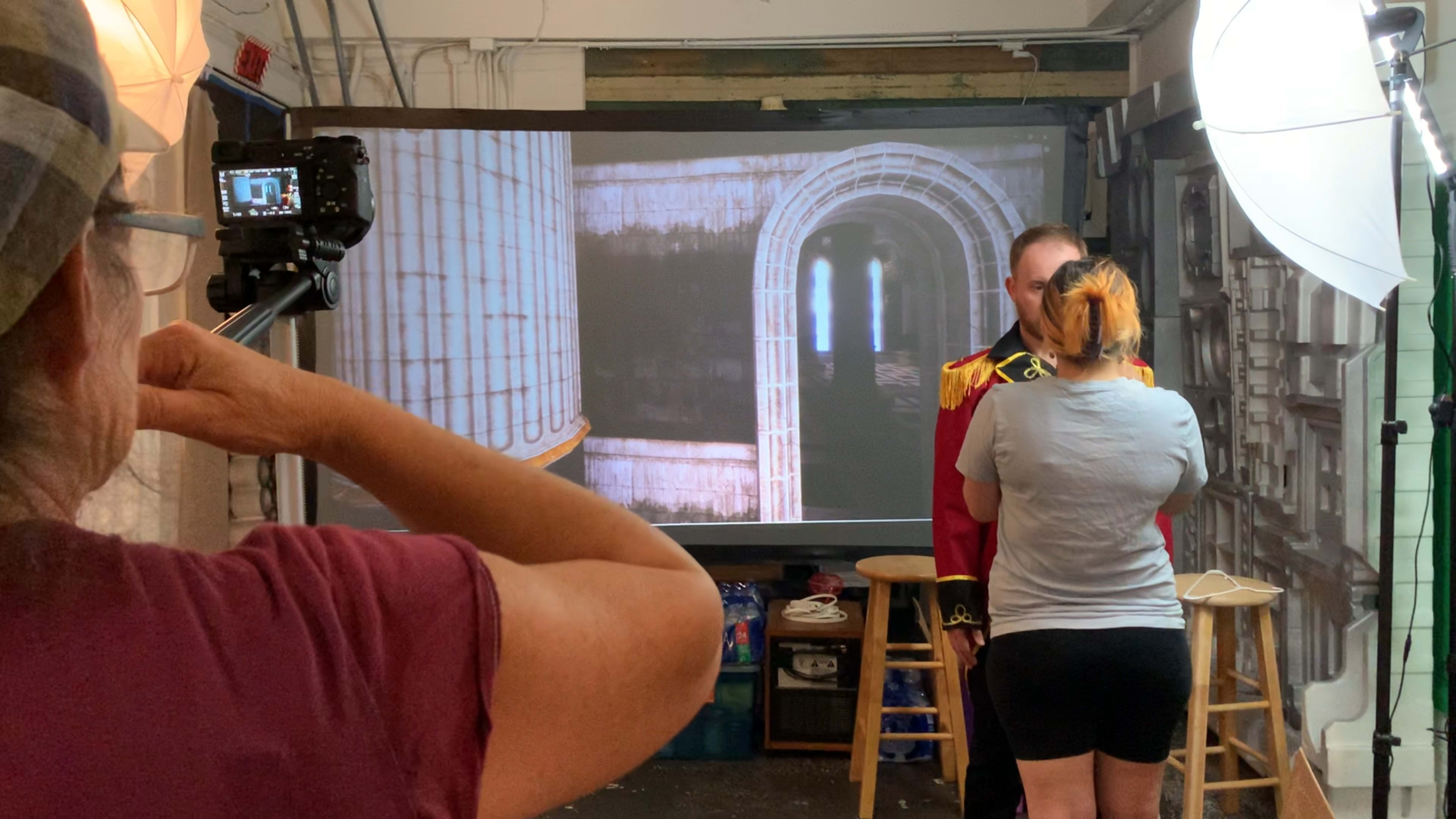 A person in a red costume stands in front of a projector displaying an architectural backdrop, while another individual adjusts equipment in the foreground.