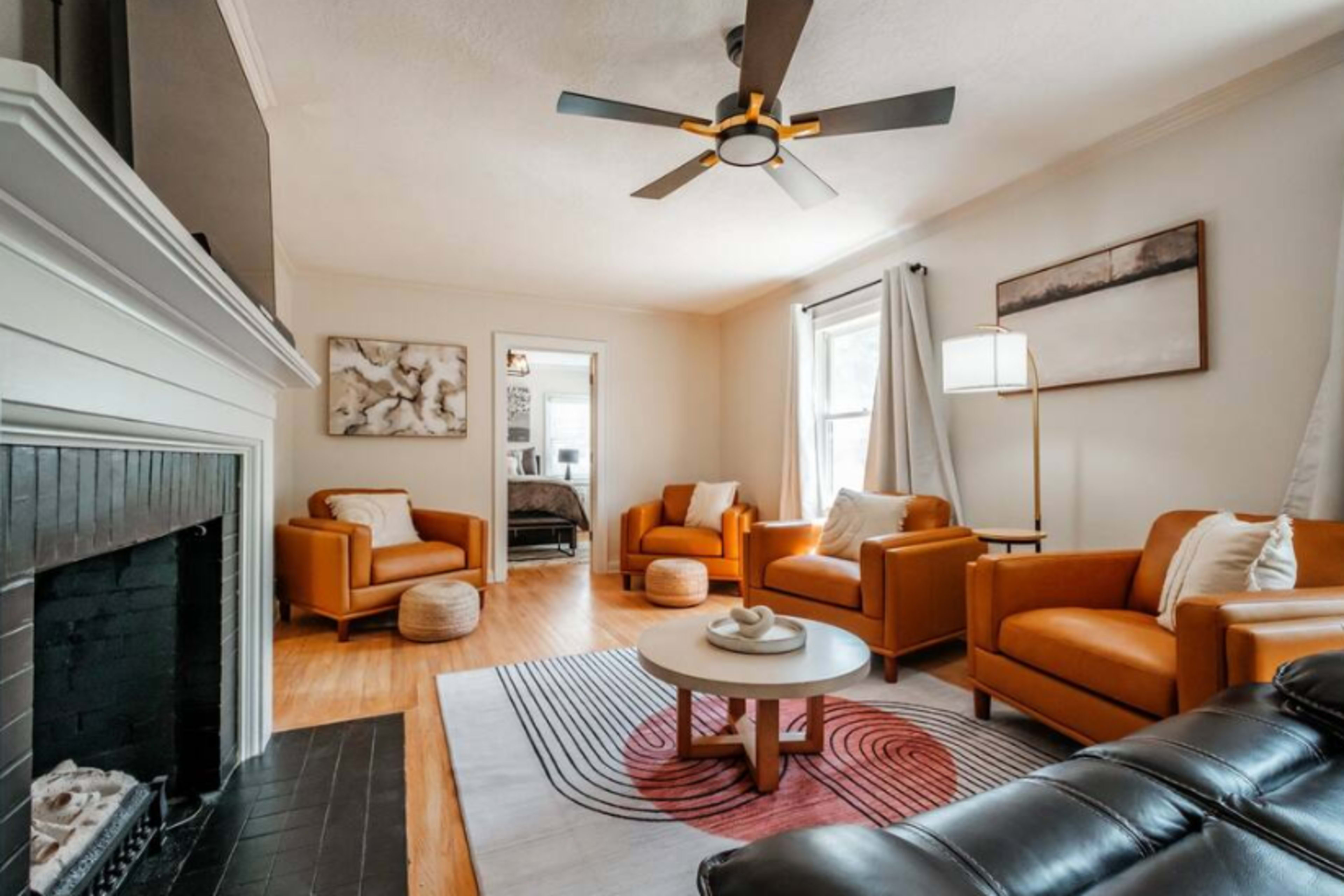 The image depicts a cozy living room featuring two orange armchairs, a black leather sofa, a round coffee table, and a ceiling fan, with a fireplace on one side and natural light coming through the windows.