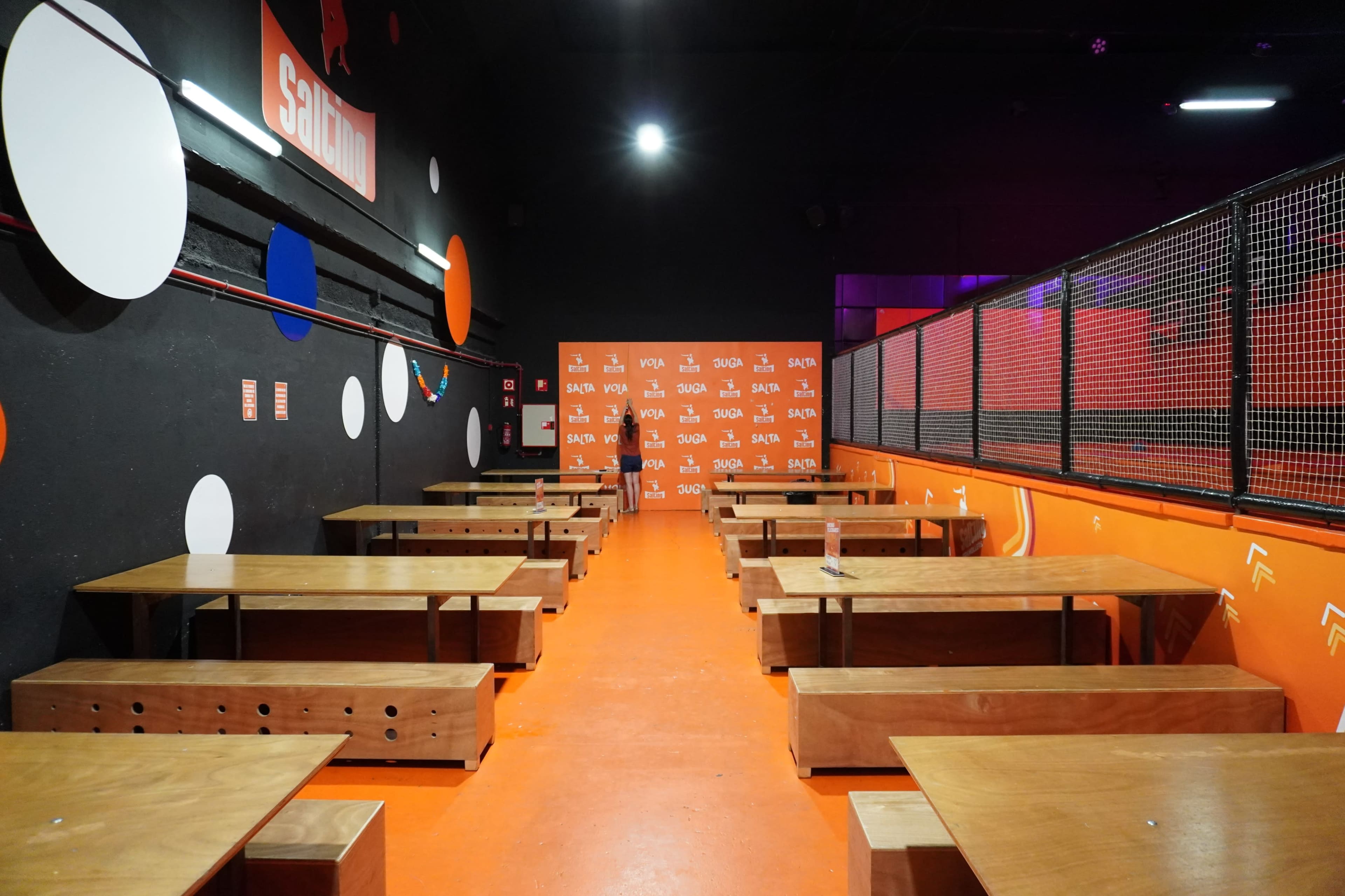 A spacious indoor area with wooden benches arranged around a bright orange floor, featuring a wall decorated with colorful circular shapes.